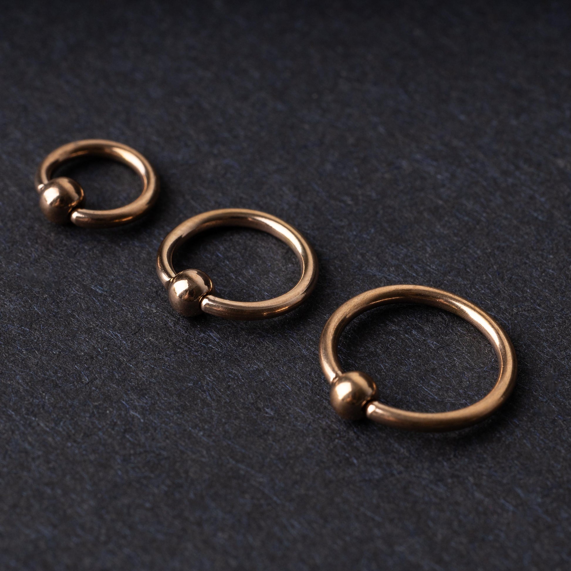 Rose Gold-tone ball closure rings in three sizes, displayed on a dark background.