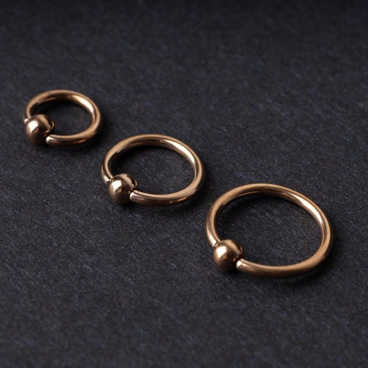 Rose Gold-tone ball closure rings in three sizes, displayed on a dark background.