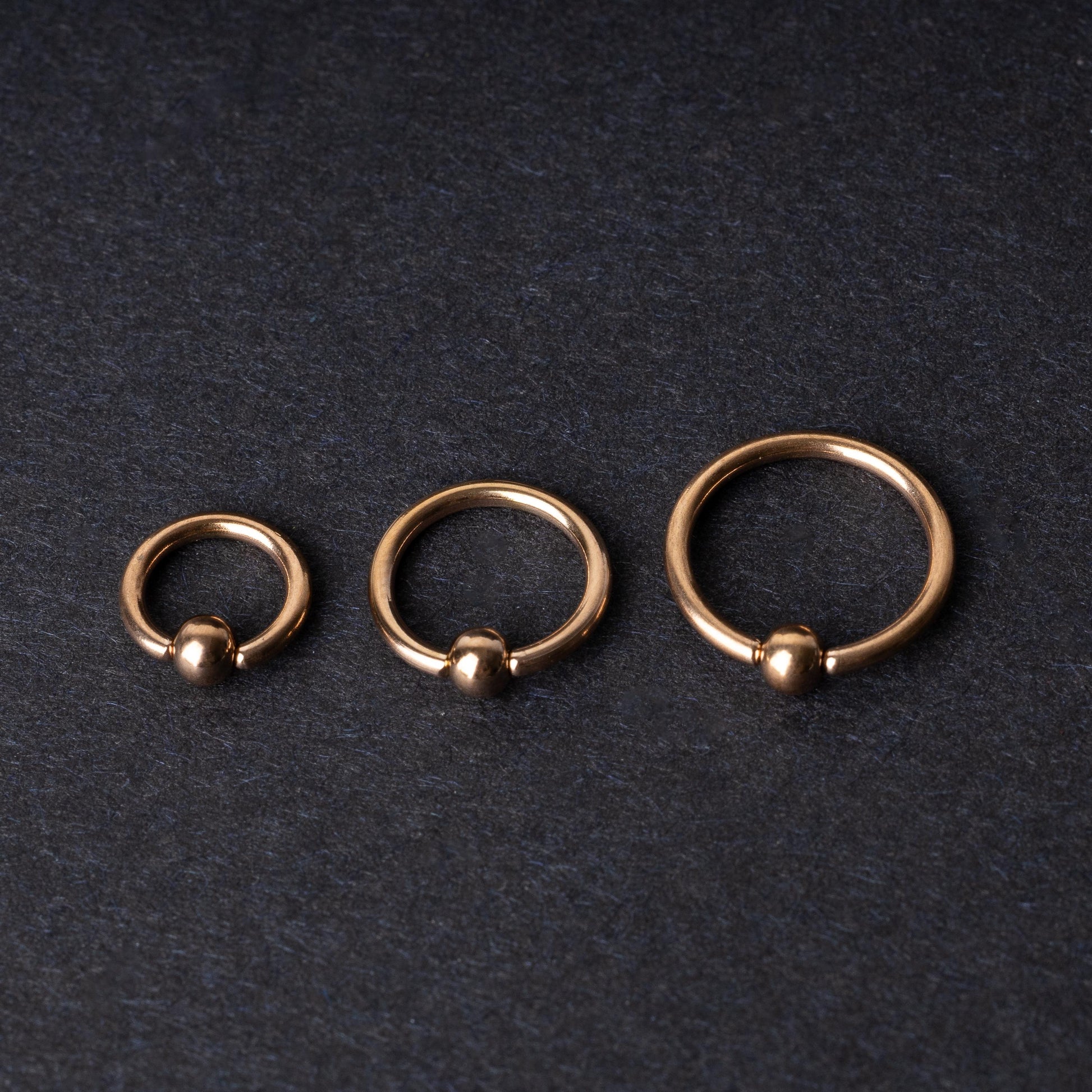 Rose Gold-tone ball closure rings in three sizes, displayed on a dark background.