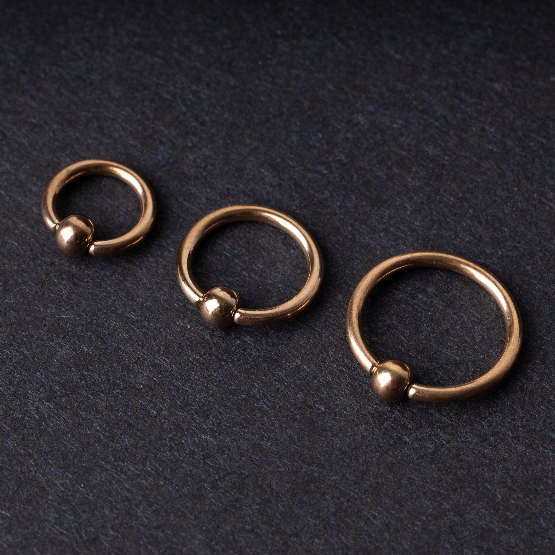 Rose Gold-tone ball closure rings in three sizes, displayed on a dark background.