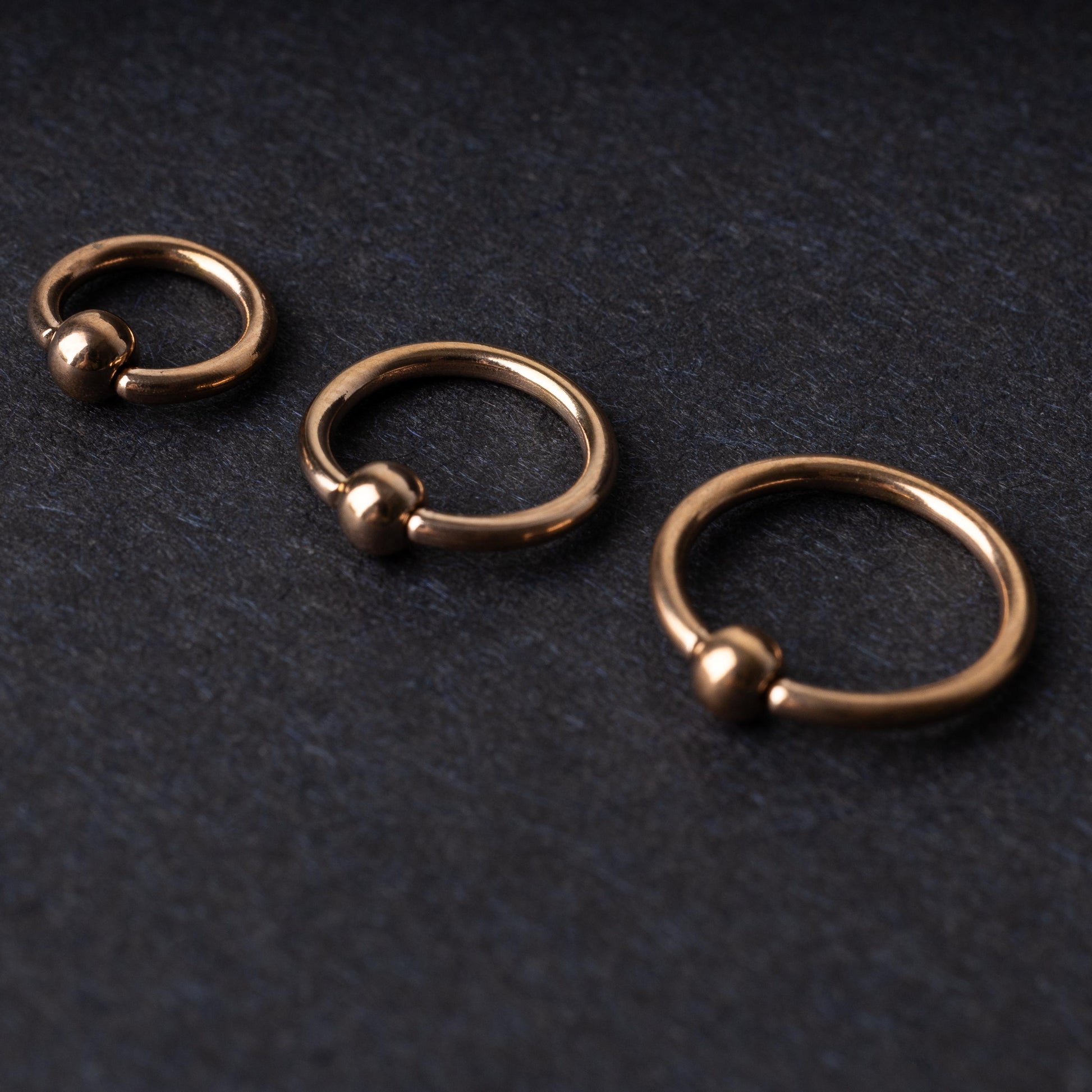 Rose Gold-tone ball closure rings in three sizes, displayed on a dark background.