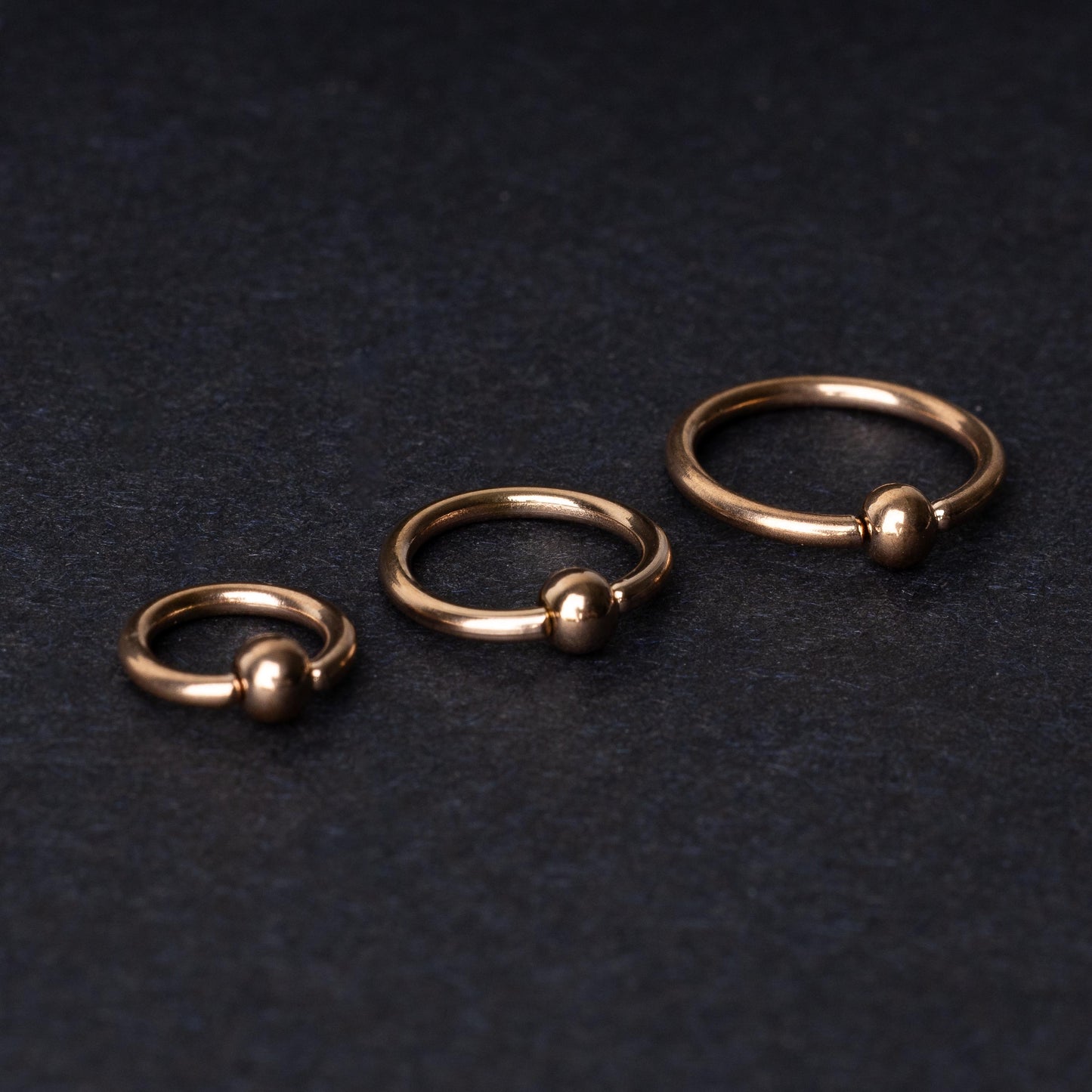 Rose Gold-tone ball closure rings in three sizes, displayed on a dark background.