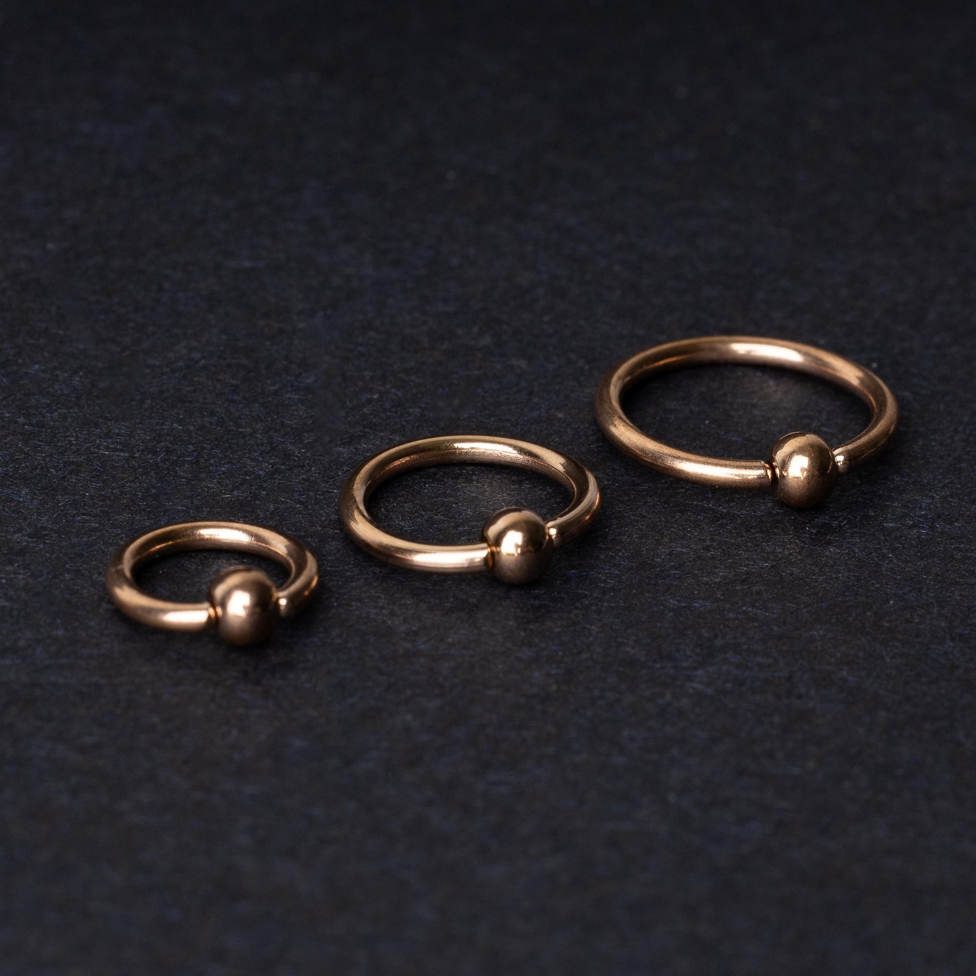 Rose Gold-tone ball closure rings in three sizes, displayed on a dark background.