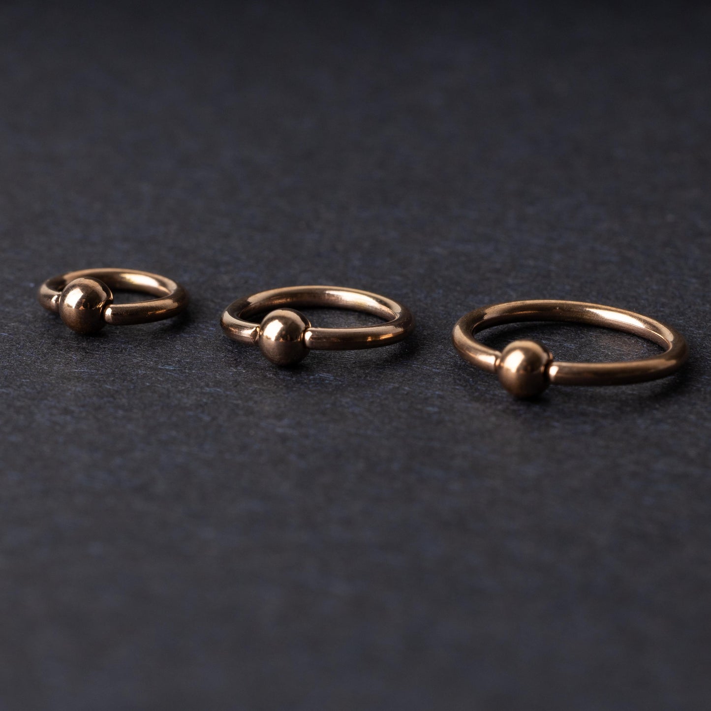 Rose Gold-tone ball closure rings in three sizes, displayed on a dark background.