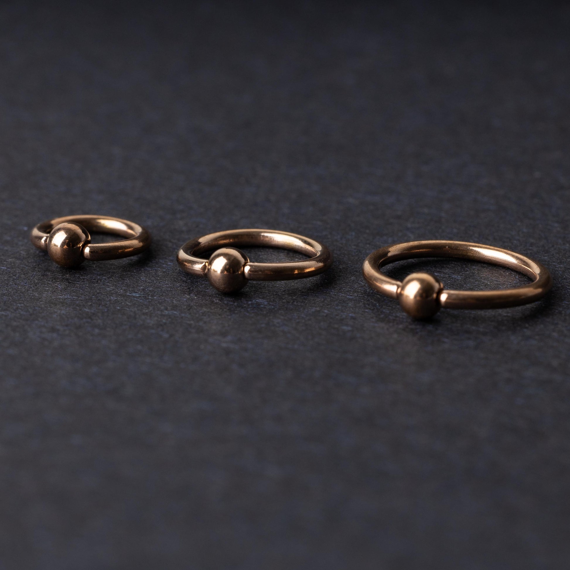 Rose Gold-tone ball closure rings in three sizes, displayed on a dark background.