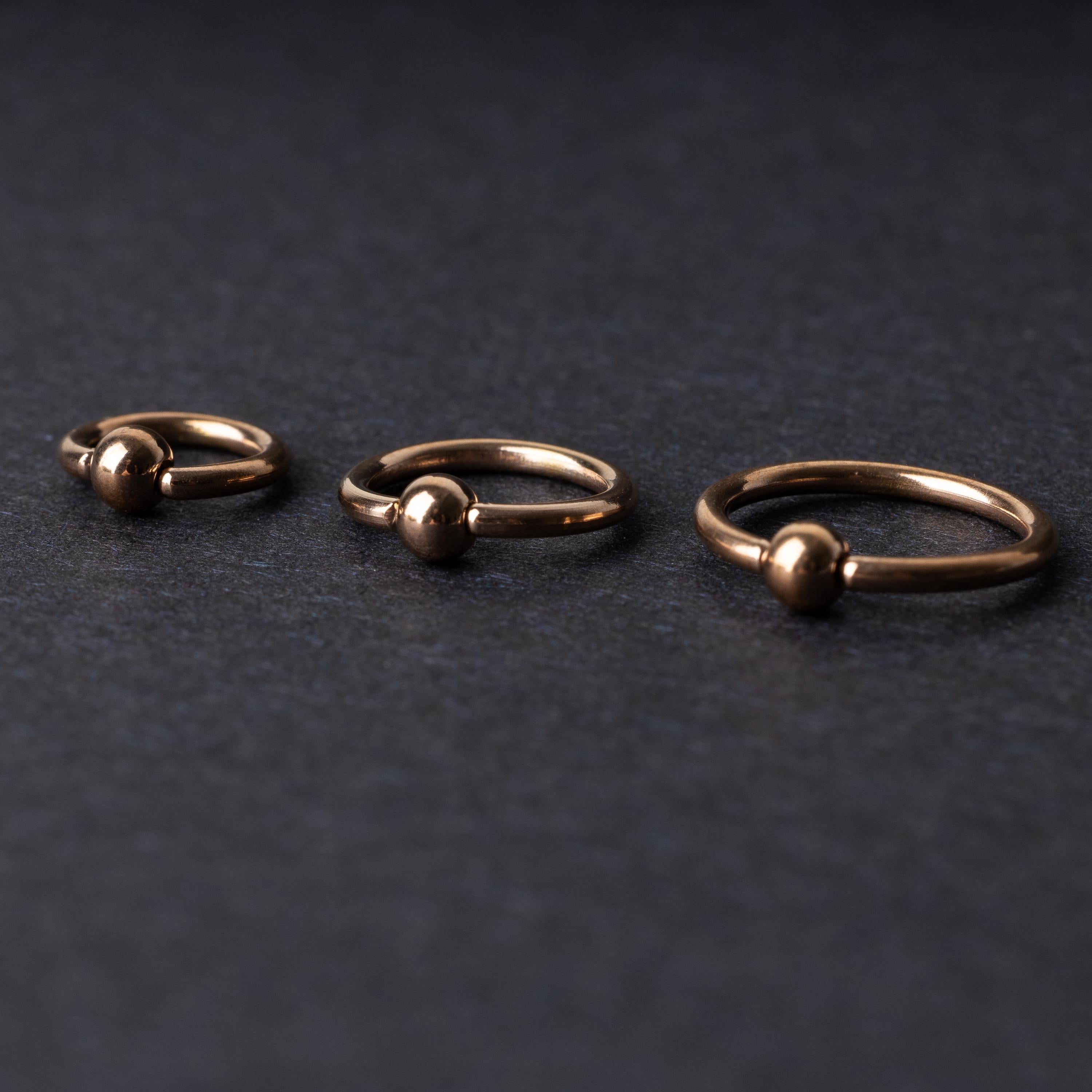 Rose Gold-tone ball closure rings in three sizes, displayed on a dark background.