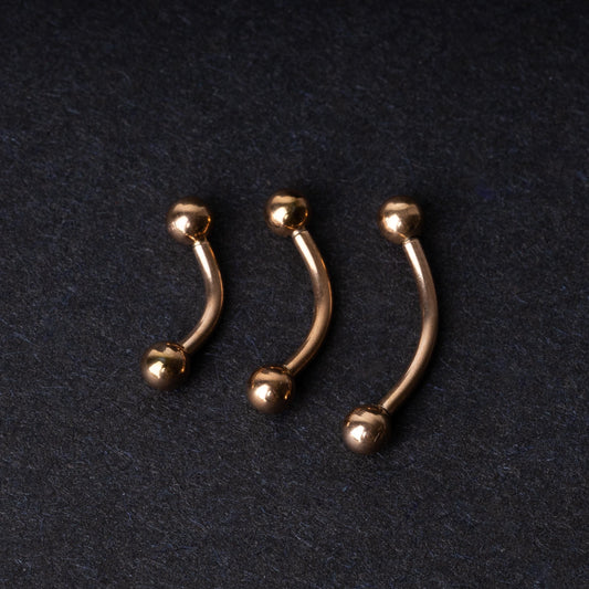Three rose gold-tone stainless steel curved barbells of varying sizes displayed on a dark background.