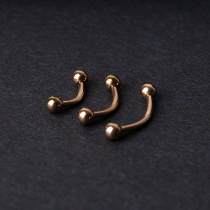 Three rose gold-tone stainless steel curved barbells of varying sizes displayed on a dark background.