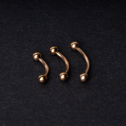 Three rose gold-tone stainless steel curved barbells of varying sizes displayed on a dark background.