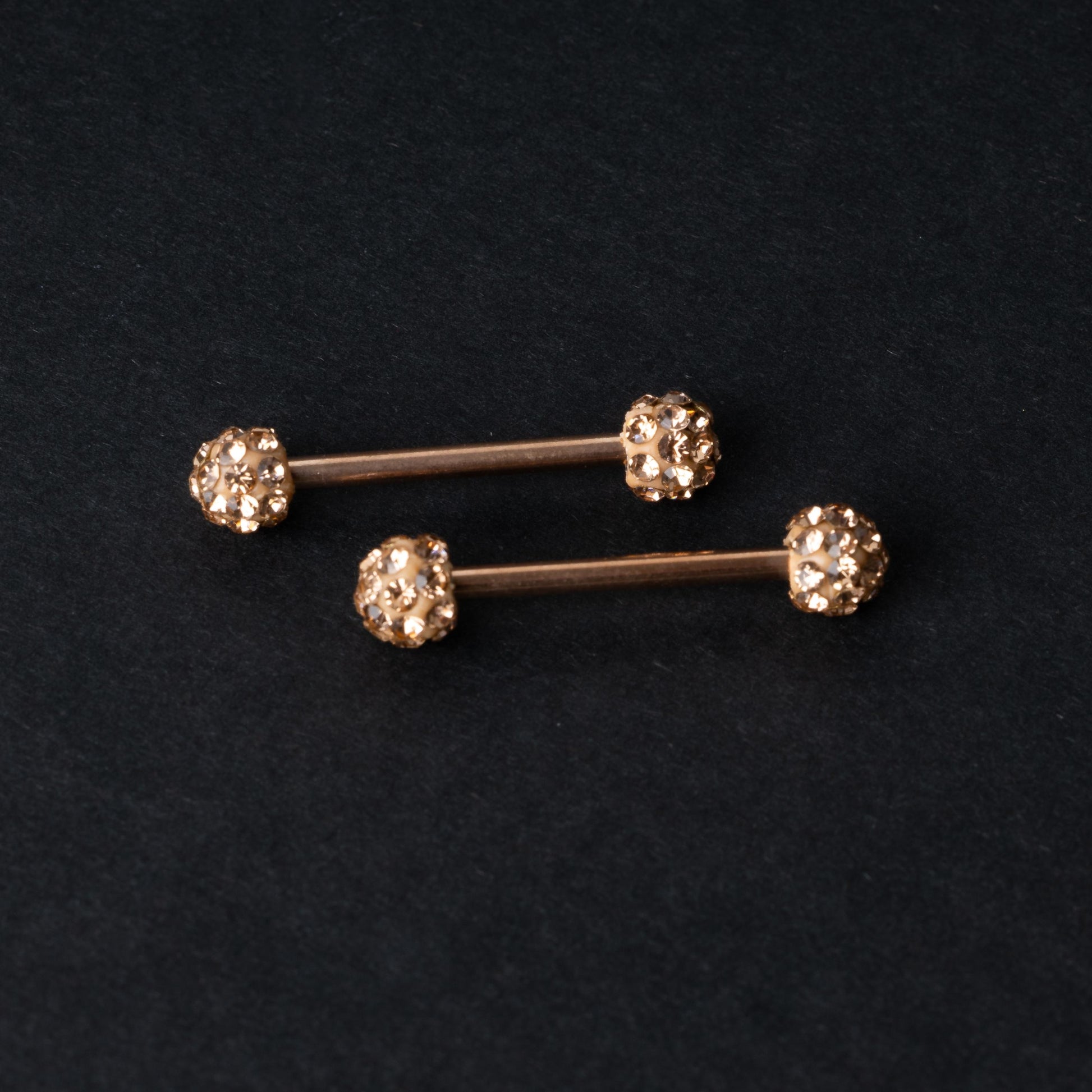 Barbells with textured crystal-encrusted ends in rose gold displayed on a dark background.