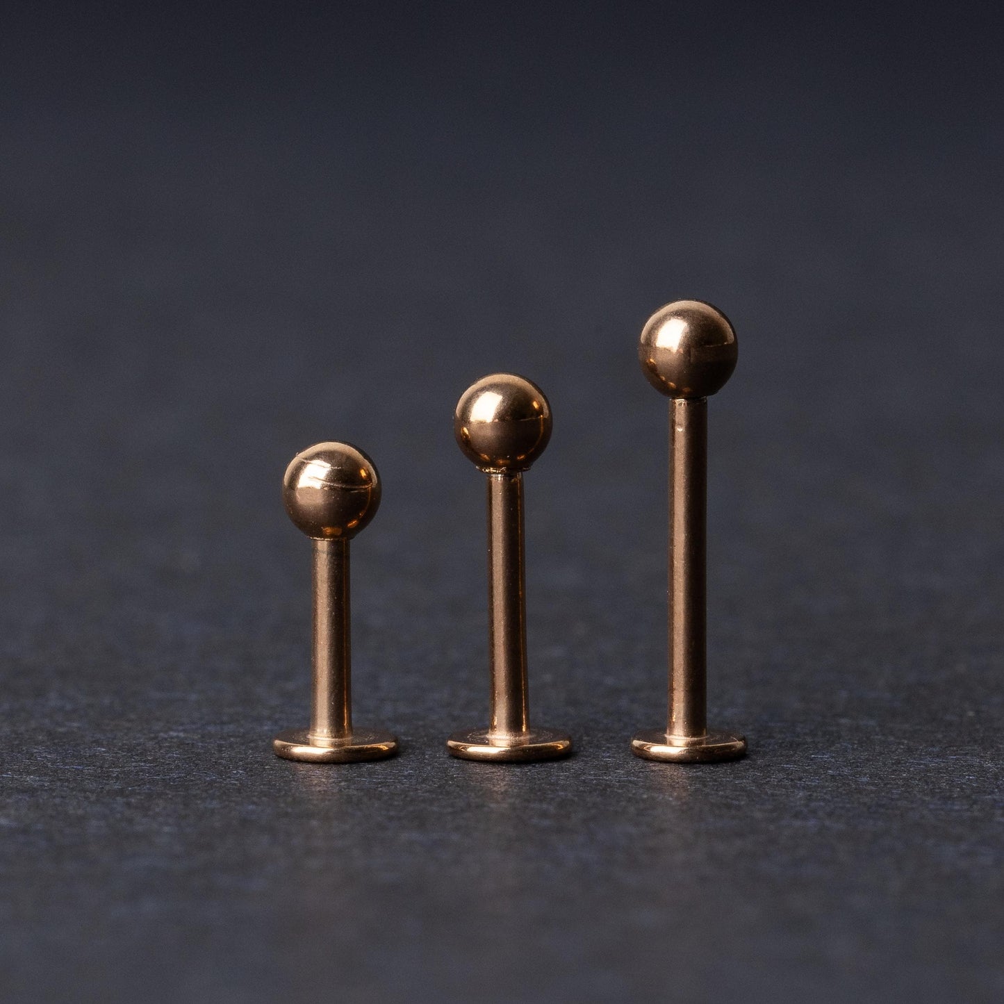 Three rose gold-tone stainless steel labret studs of varying lengths, featuring smooth ball ends, displayed on a dark background.