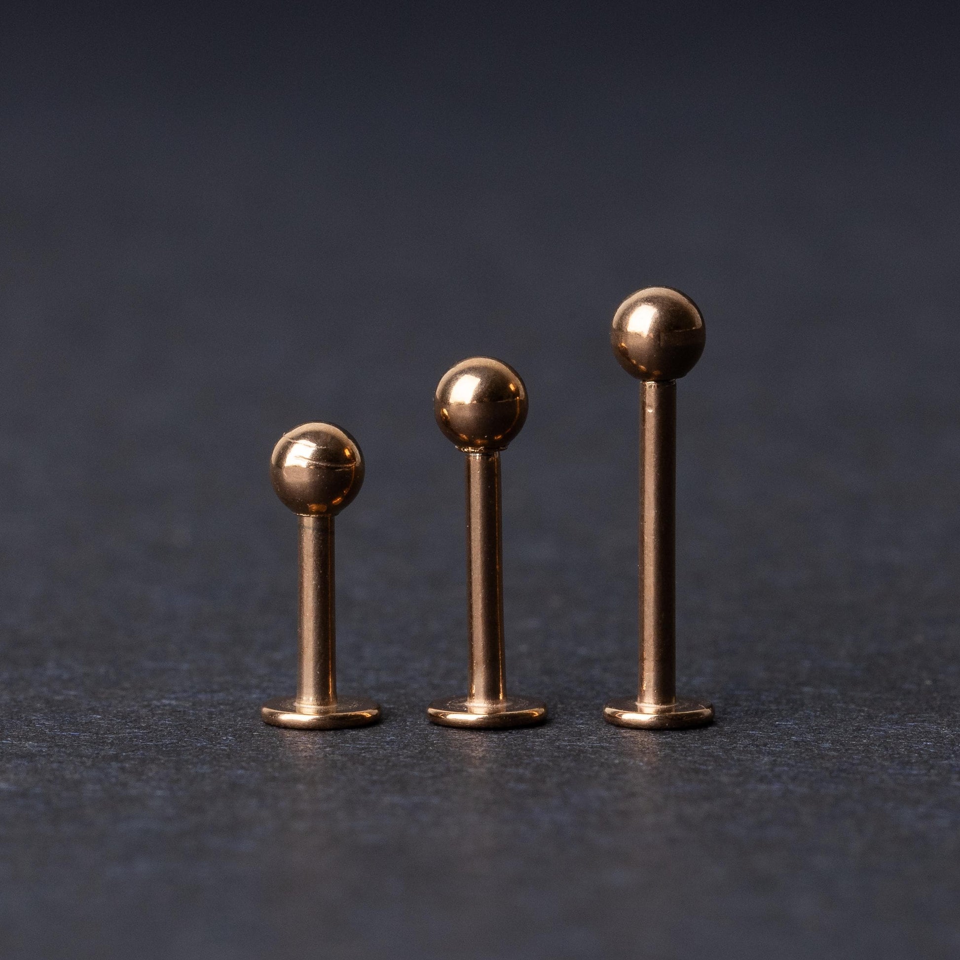 Three rose gold-tone stainless steel labret studs of varying lengths, featuring smooth ball ends, displayed on a dark background.