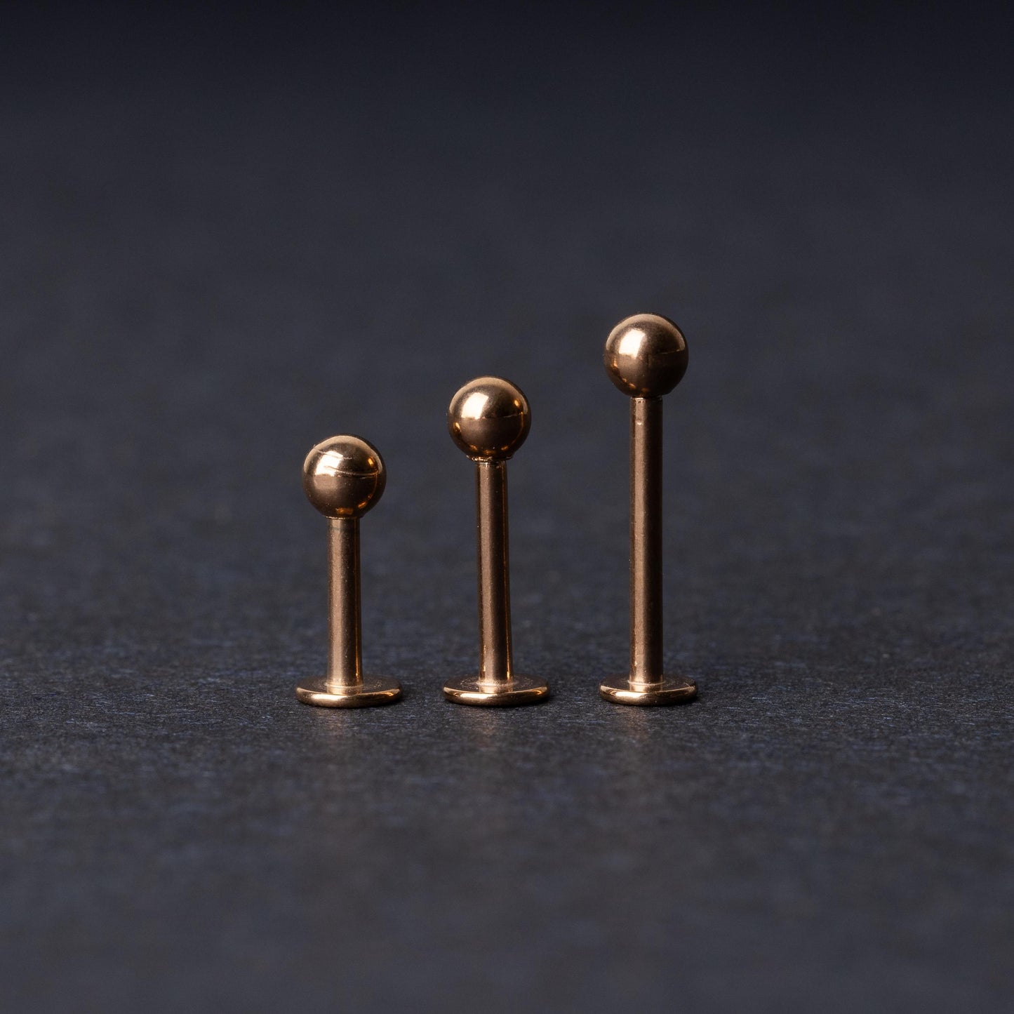 Three rose gold-tone stainless steel labret studs of varying lengths, featuring smooth ball ends, displayed on a dark background.