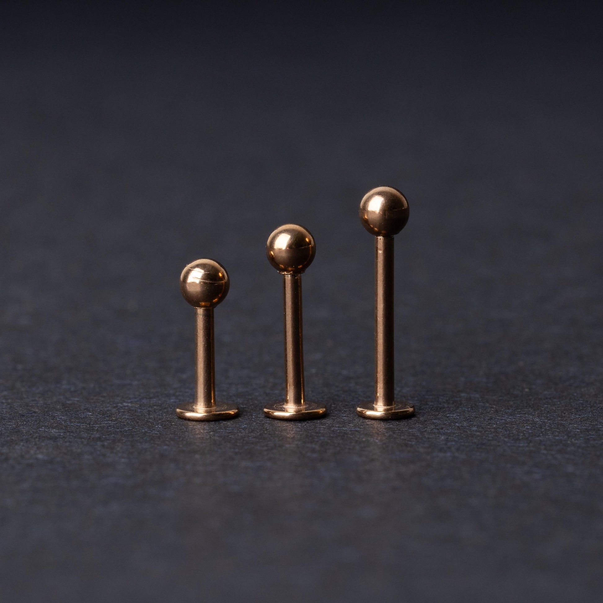 Three rose gold-tone stainless steel labret studs of varying lengths, featuring smooth ball ends, displayed on a dark background.