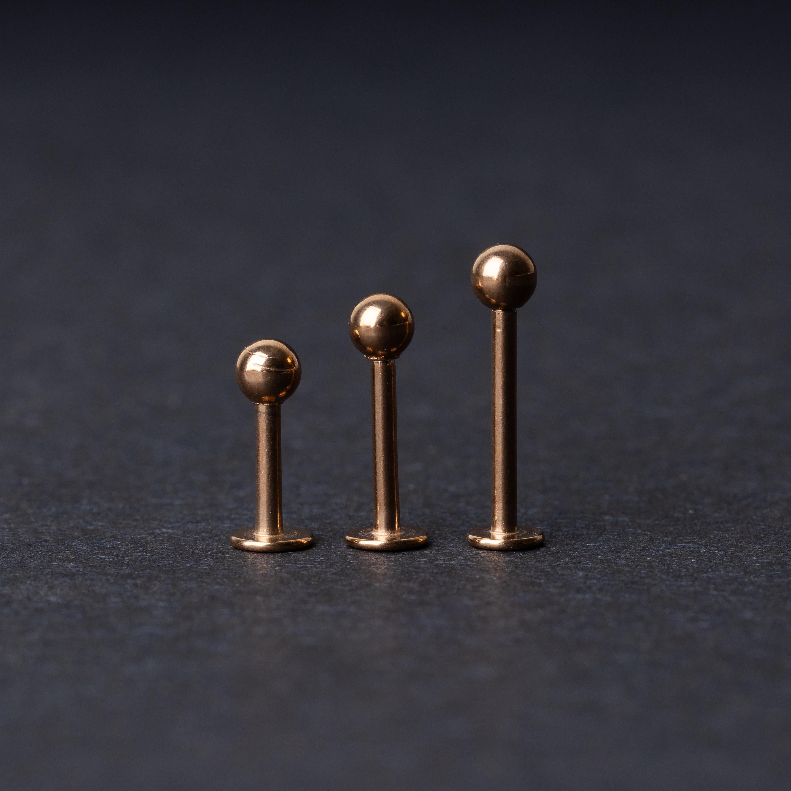 Three rose gold-tone stainless steel labret studs of varying lengths, featuring smooth ball ends, displayed on a dark background.
