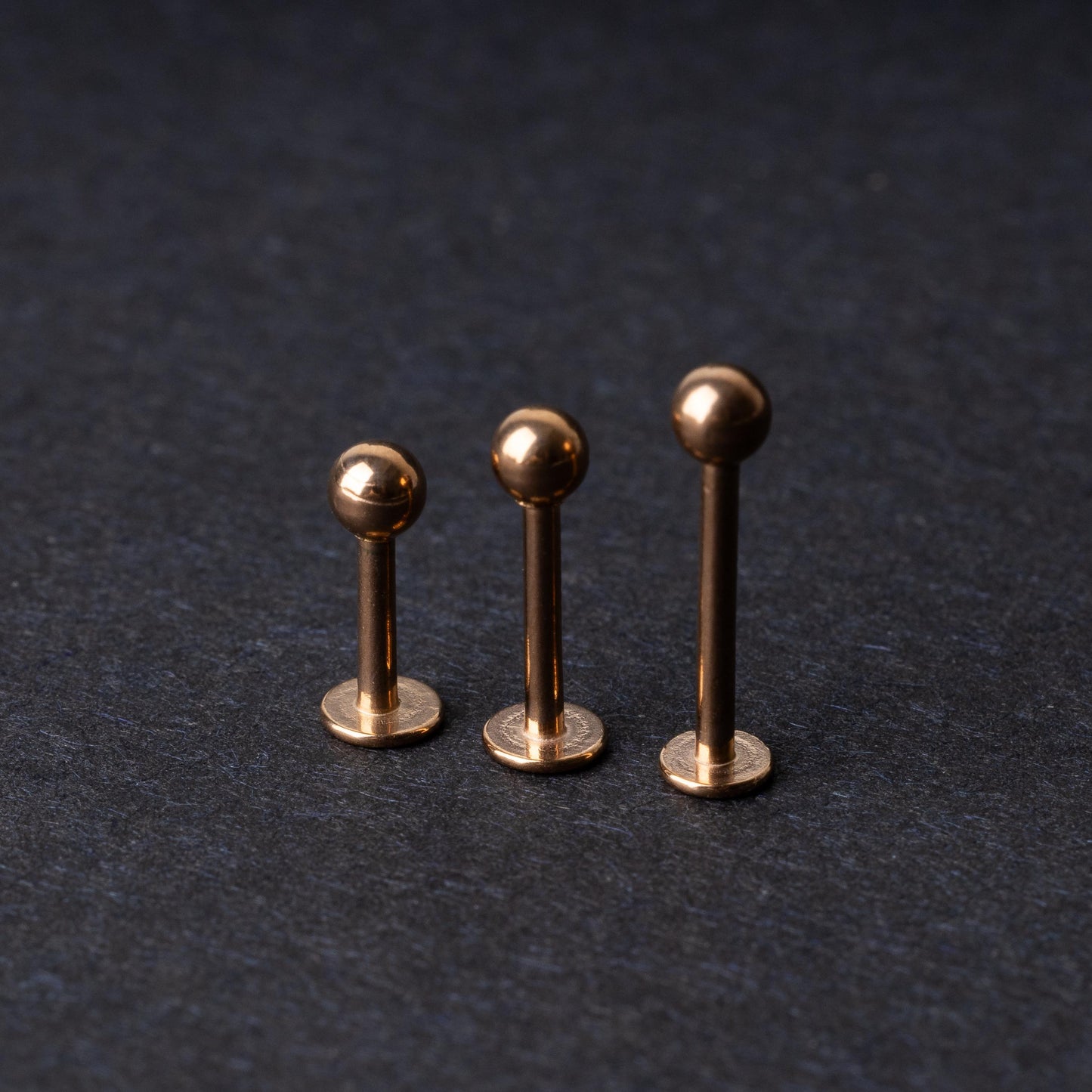 Three rose gold-tone stainless steel labret studs of varying lengths, featuring smooth ball ends, displayed on a dark background.