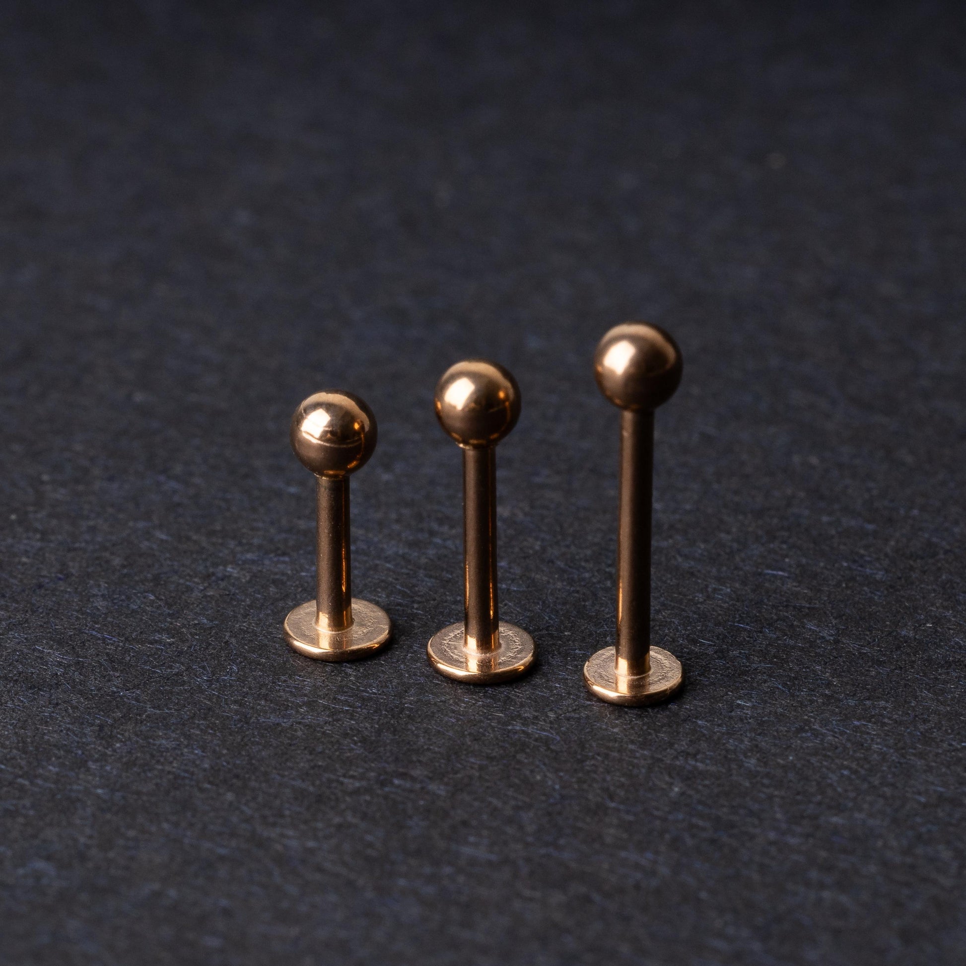 Three rose gold-tone stainless steel labret studs of varying lengths, featuring smooth ball ends, displayed on a dark background.