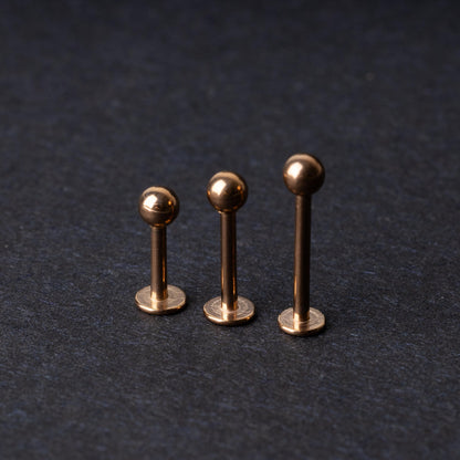 Three rose gold-tone stainless steel labret studs of varying lengths, featuring smooth ball ends, displayed on a dark background.