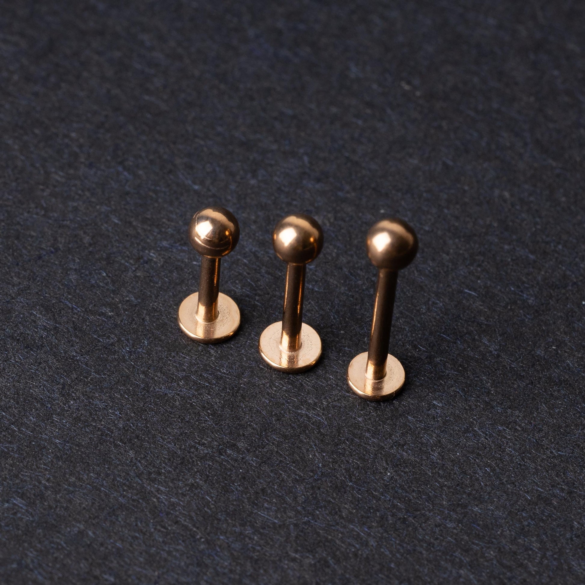 Three rose gold-tone stainless steel labret studs of varying lengths, featuring smooth ball ends, displayed on a dark background.