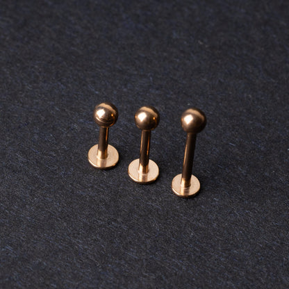 Three rose gold-tone stainless steel labret studs of varying lengths, featuring smooth ball ends, displayed on a dark background.