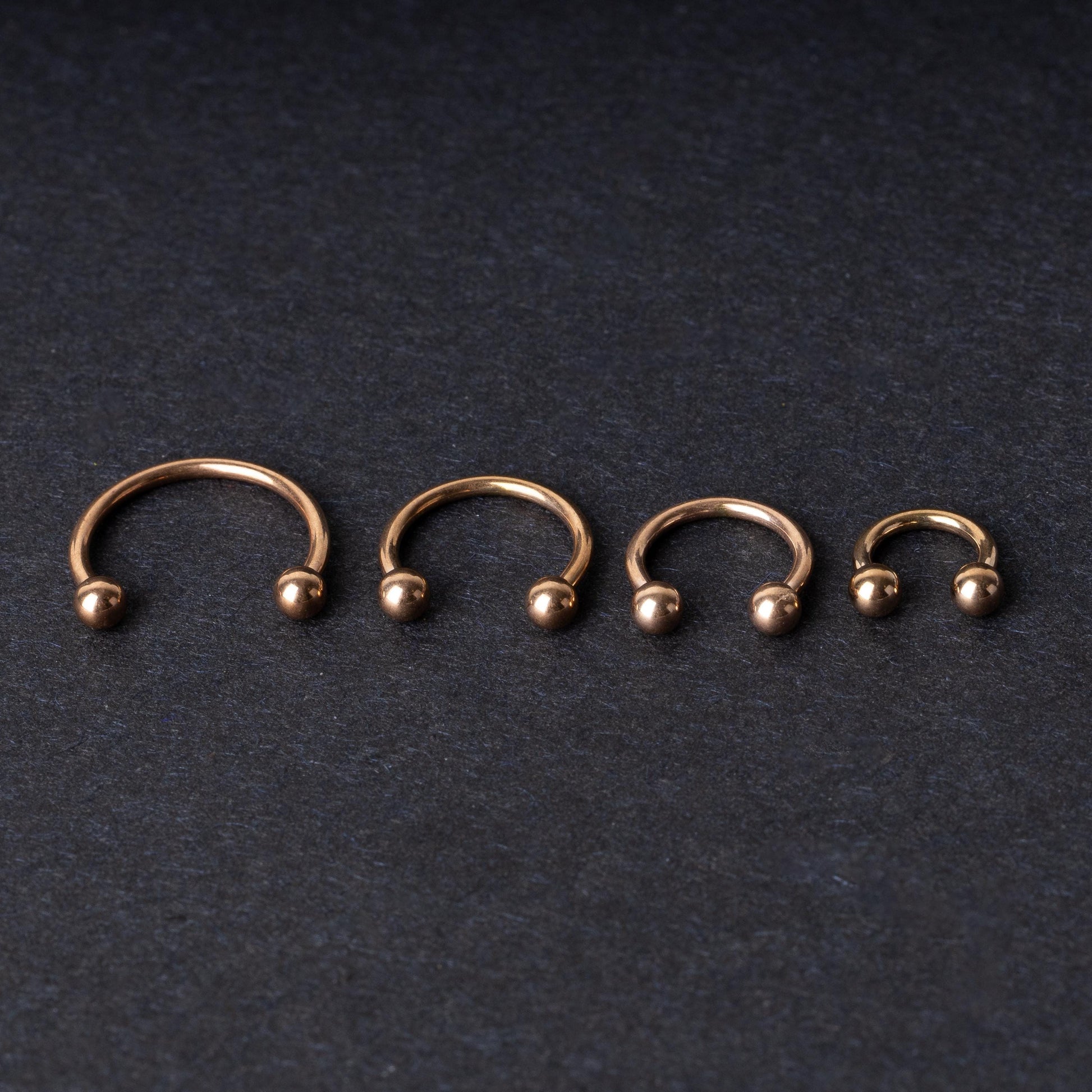 Rose Gold-tone horseshoe rings displayed in a row on a dark background, showcasing various sizes for septum or body piercings.
