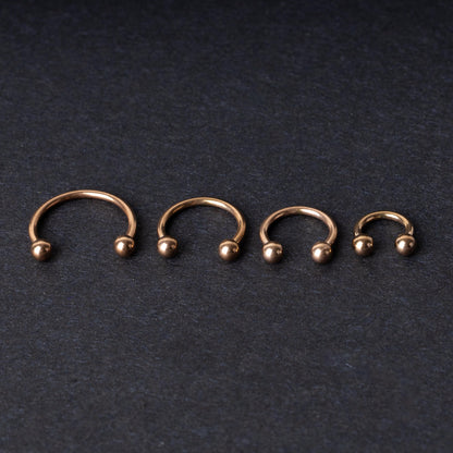 Rose Gold-tone horseshoe rings displayed in a row on a dark background, showcasing various sizes for septum or body piercings.
