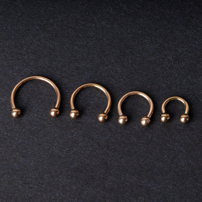 Rose Gold-tone horseshoe rings displayed in a row on a dark background, showcasing various sizes for septum or body piercings.
