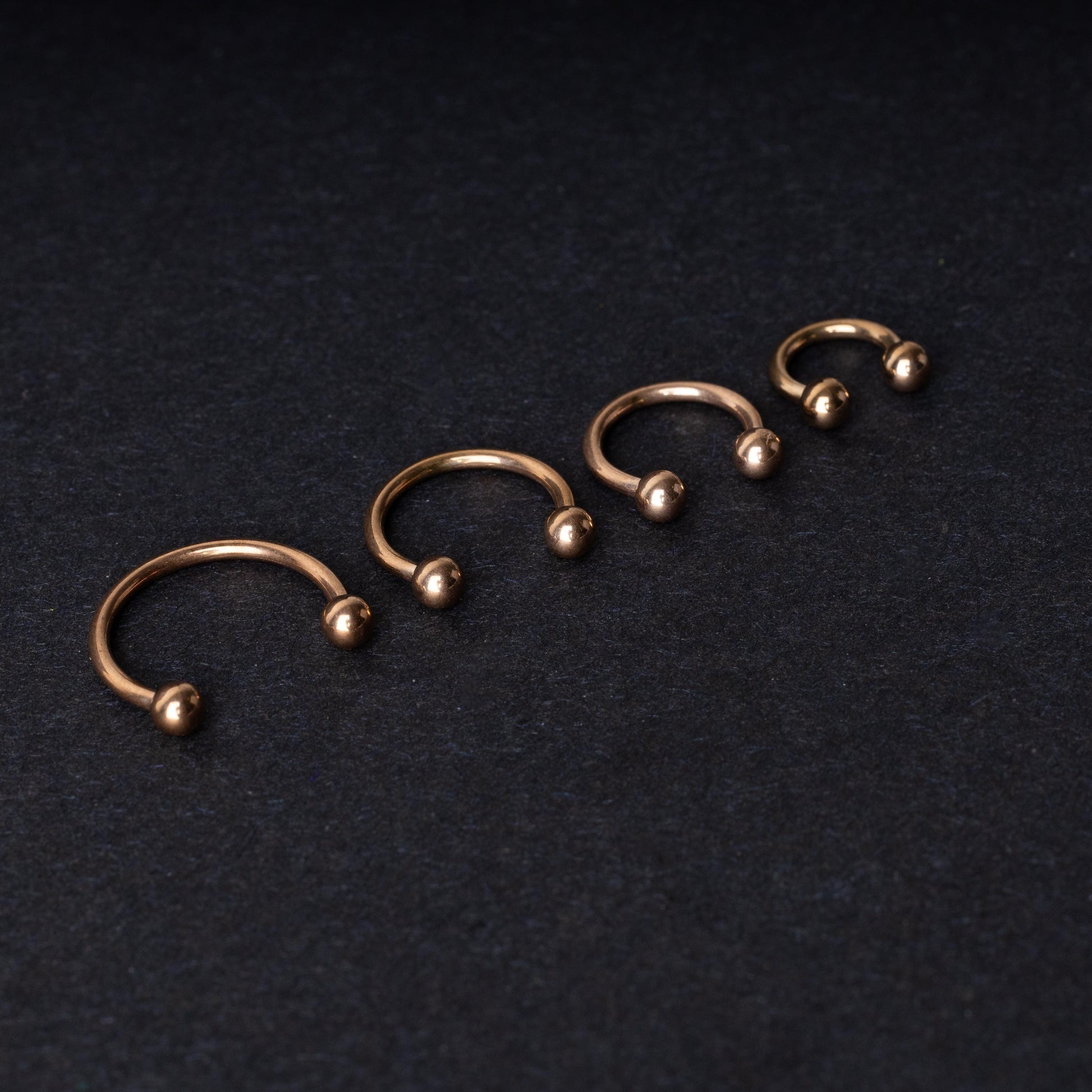 Rose Gold-tone horseshoe rings displayed in a row on a dark background, showcasing various sizes for septum or body piercings.
