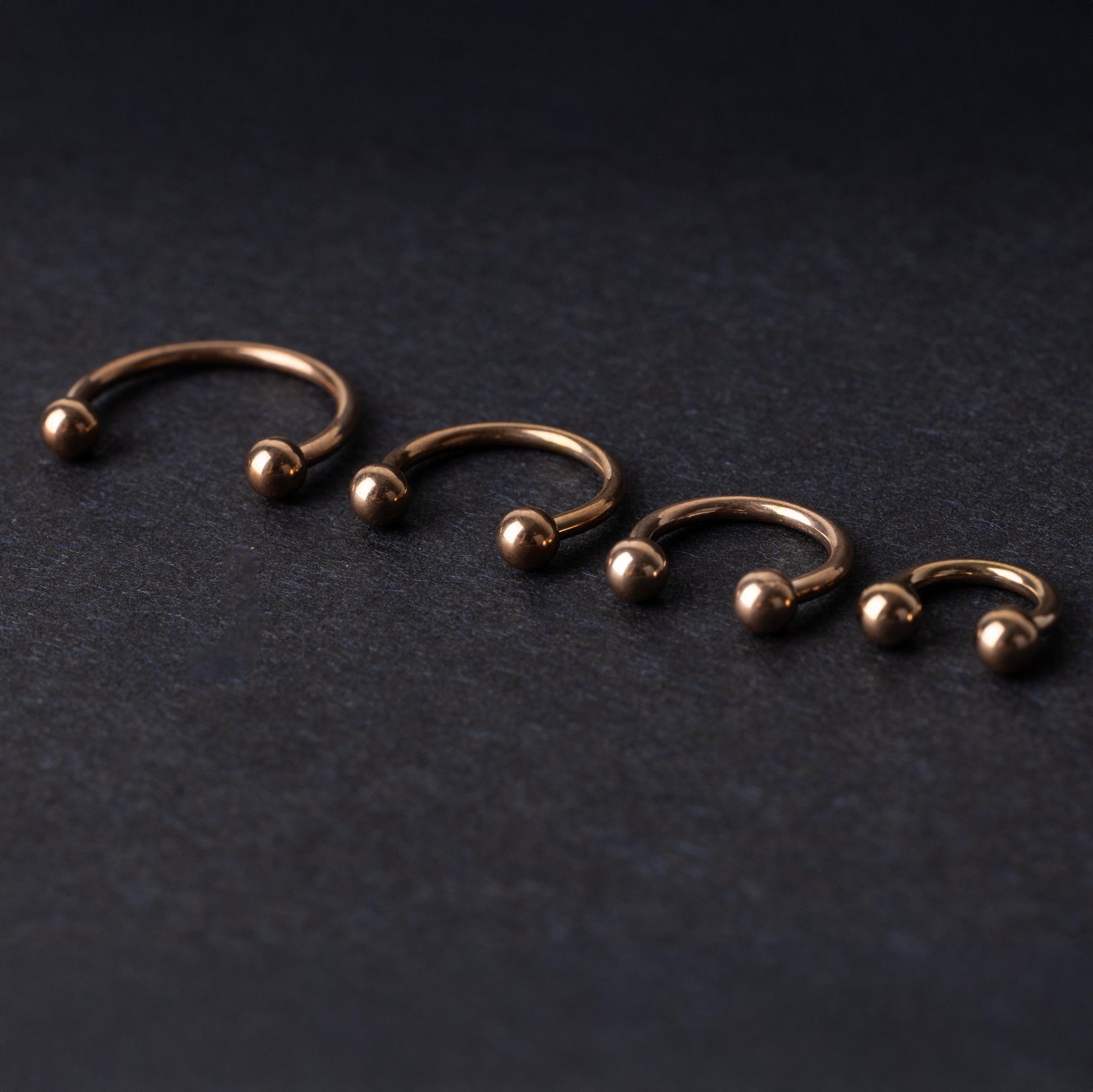 Rose Gold-tone horseshoe rings displayed in a row on a dark background, showcasing various sizes for septum or body piercings.
