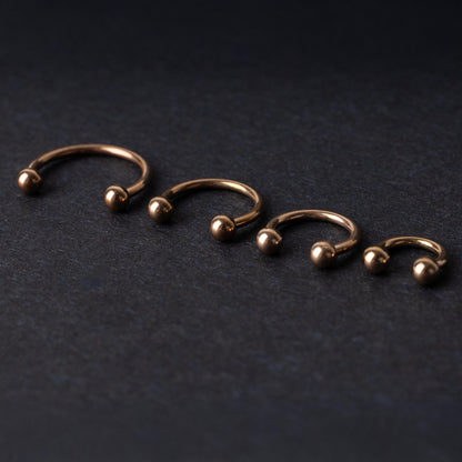 Rose Gold-tone horseshoe rings displayed in a row on a dark background, showcasing various sizes for septum or body piercings.
