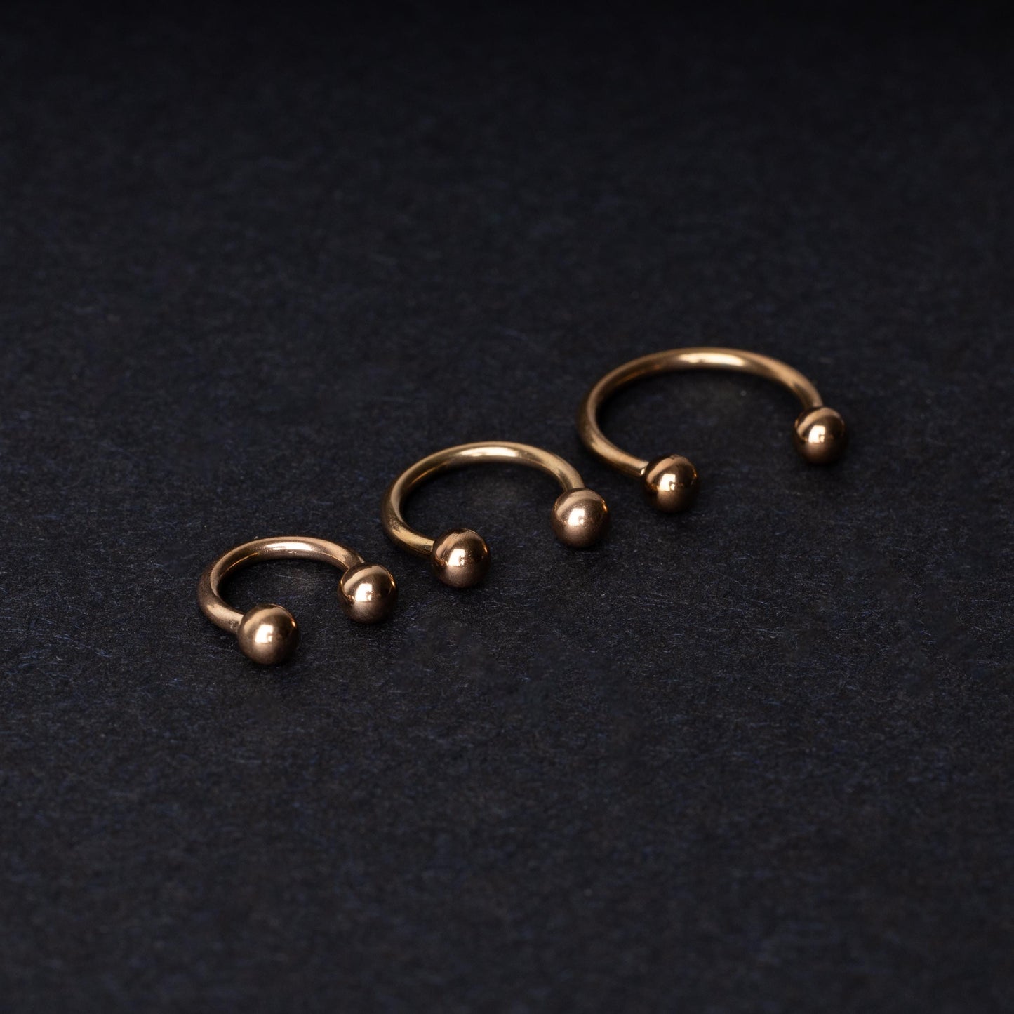 Three rose gold-tone stainless steel horseshoe hoops in varying sizes with screw-on ball ends, displayed against a dark background.