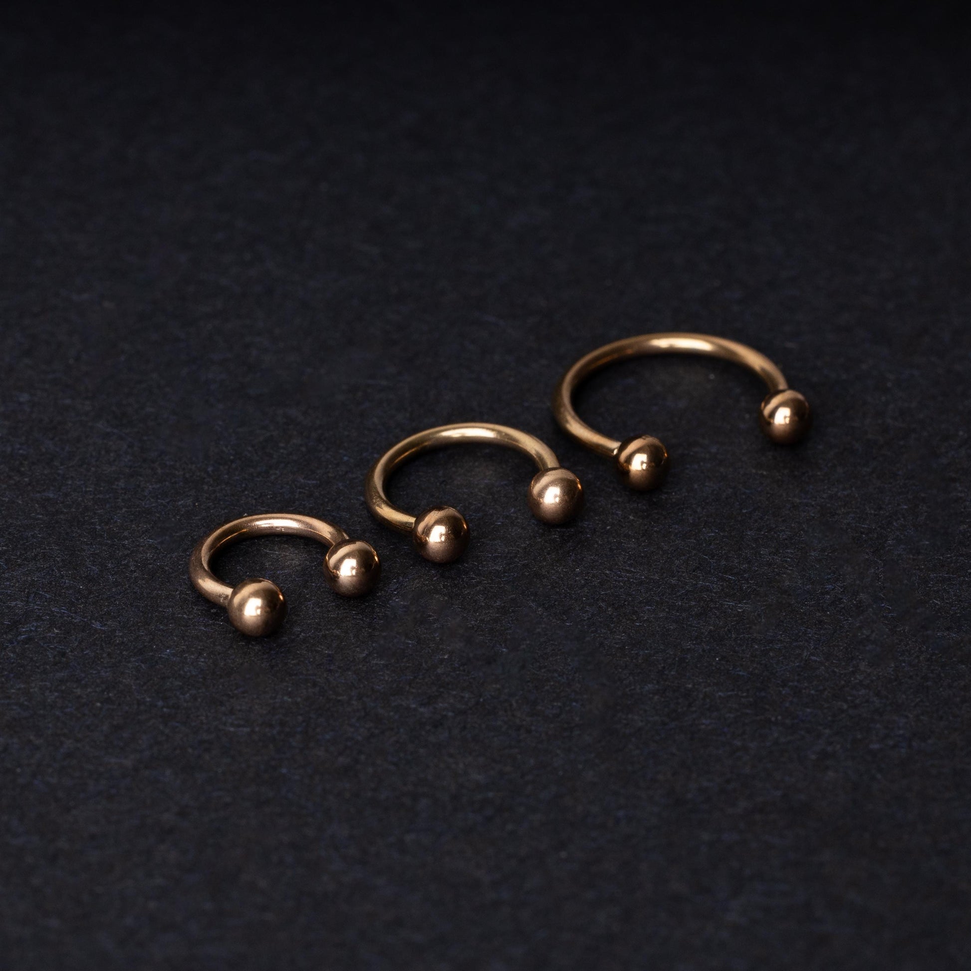 Three rose gold-tone stainless steel horseshoe hoops in varying sizes with screw-on ball ends, displayed against a dark background.
