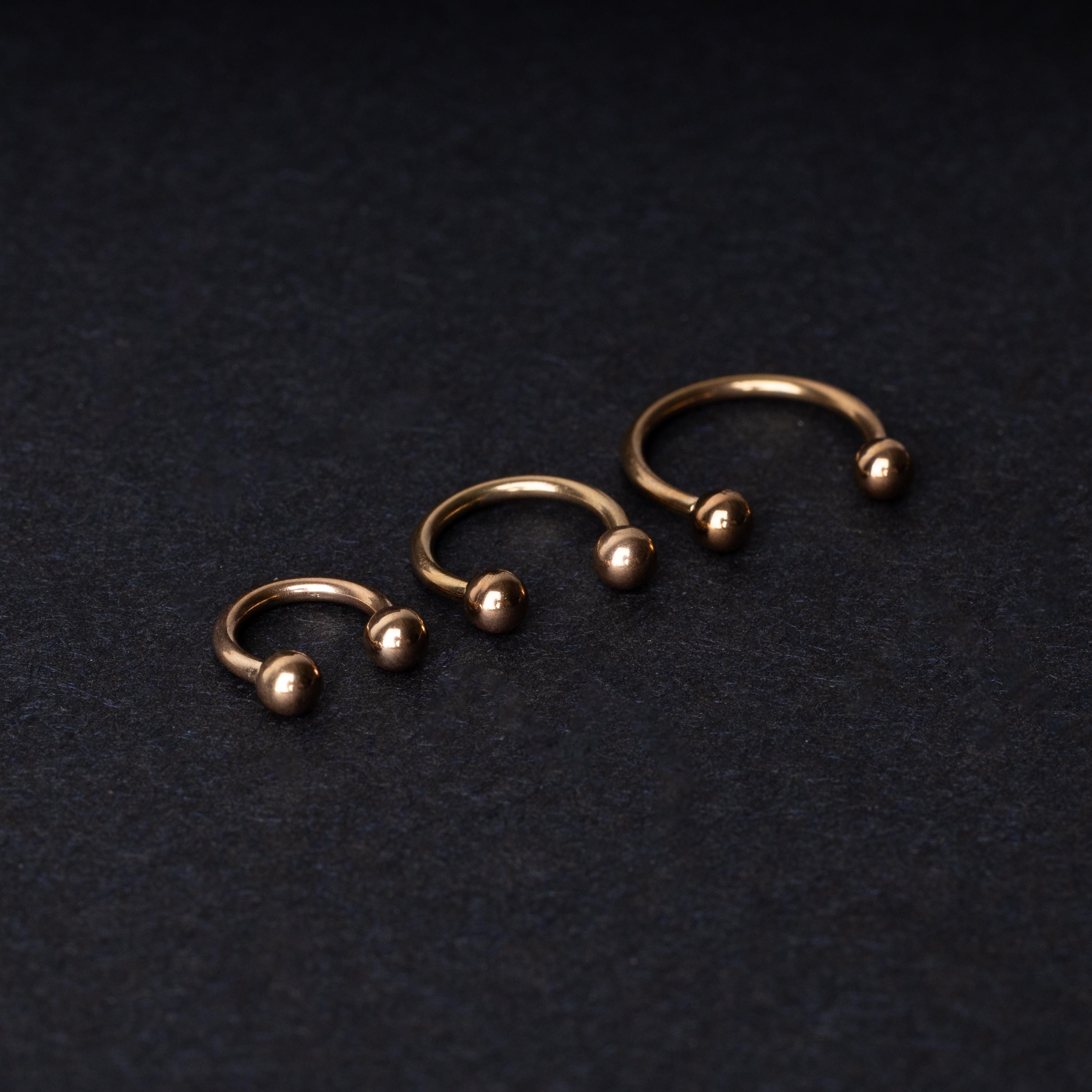 Three rose gold-tone stainless steel horseshoe hoops in varying sizes with screw-on ball ends, displayed against a dark background.