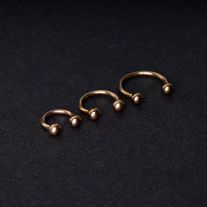 Three rose gold-tone stainless steel horseshoe hoops in varying sizes with screw-on ball ends, displayed against a dark background.