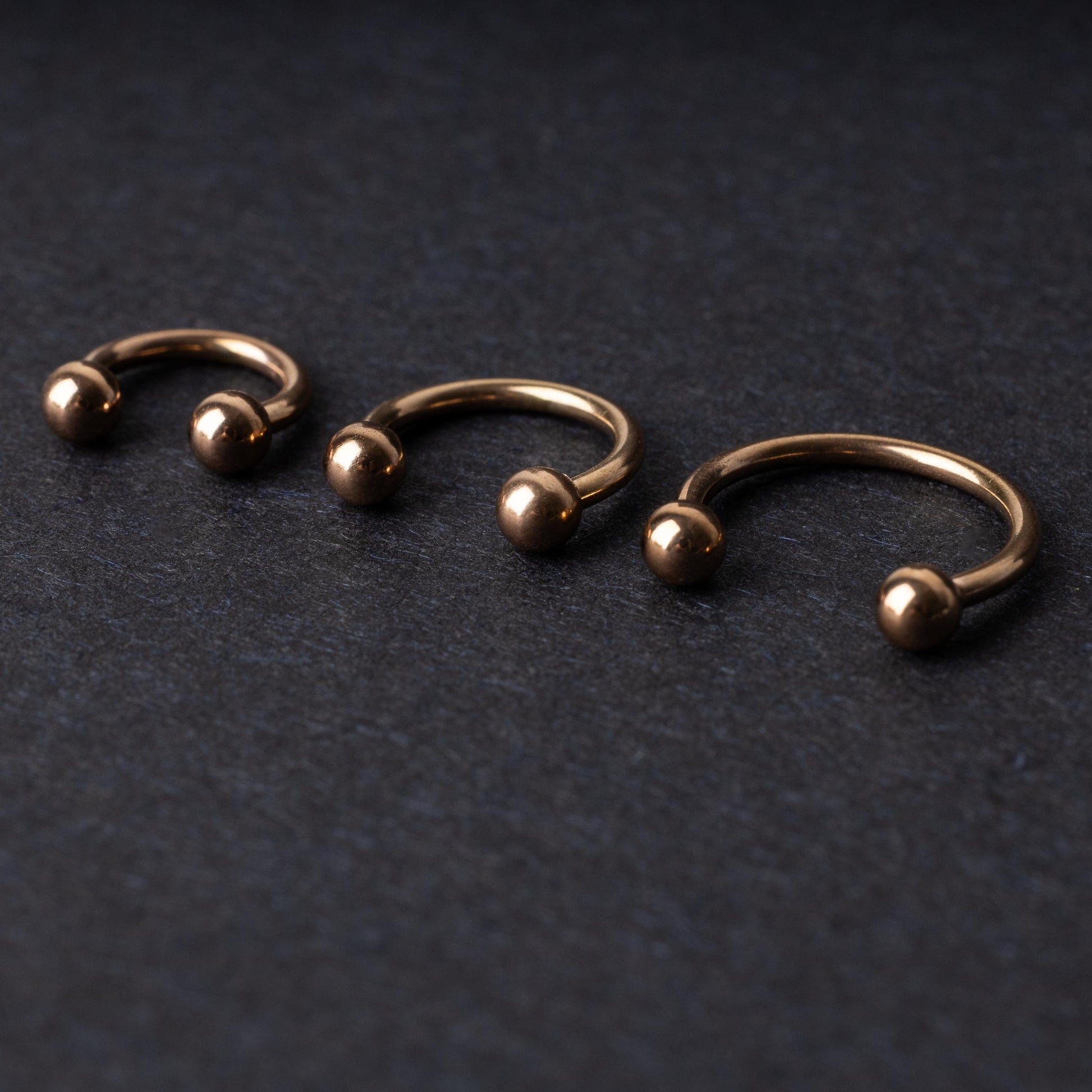 Three rose gold-tone stainless steel horseshoe hoops in varying sizes with screw-on ball ends, displayed against a dark background.