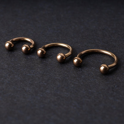 Three rose gold-tone stainless steel horseshoe hoops in varying sizes with screw-on ball ends, displayed against a dark background.