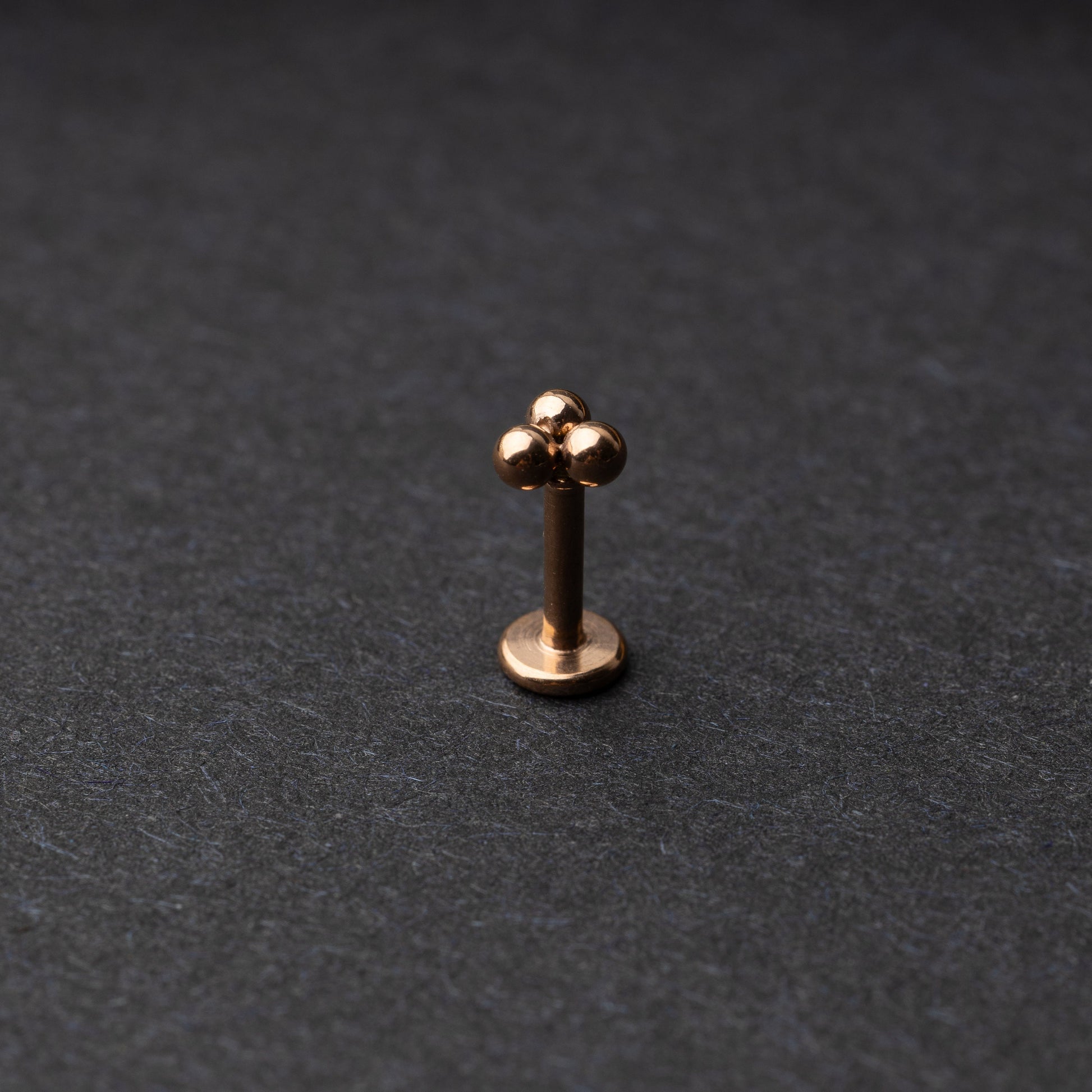 Close-up of three minimalist ball cluster labret studs in a rose gold finish, displayed on a dark background.
