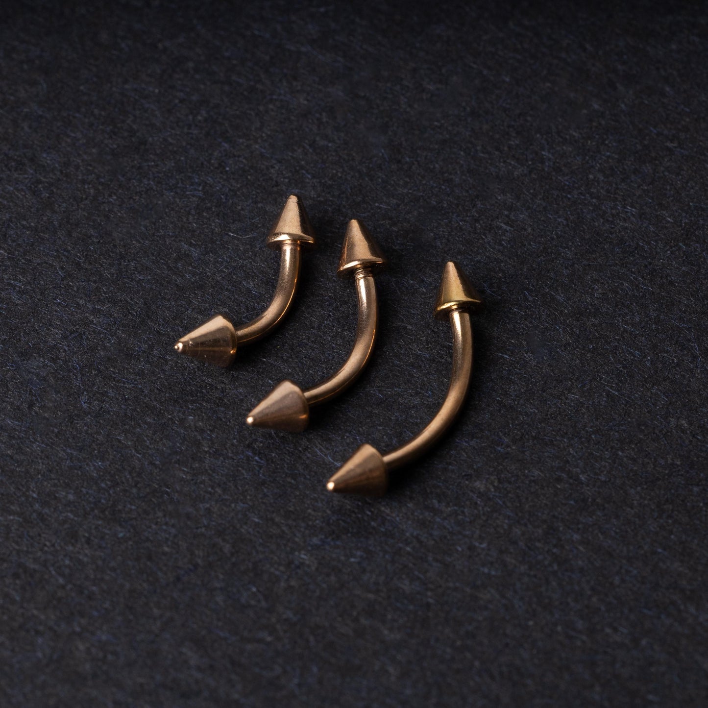 Three gold-tone titanium curved barbells with cone ends, displayed on a dark textured background, showcasing their sleek curved design and polished finis