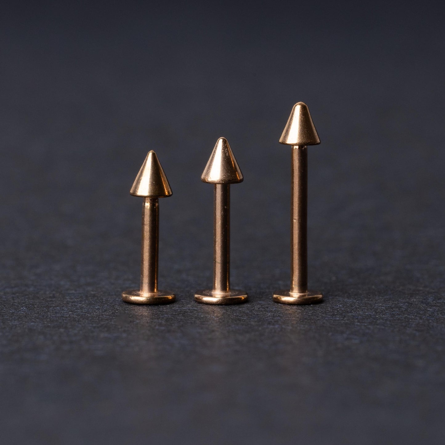 Three rose gold-tone stainless steel labrets with spike ends, standing upright on a dark background.
