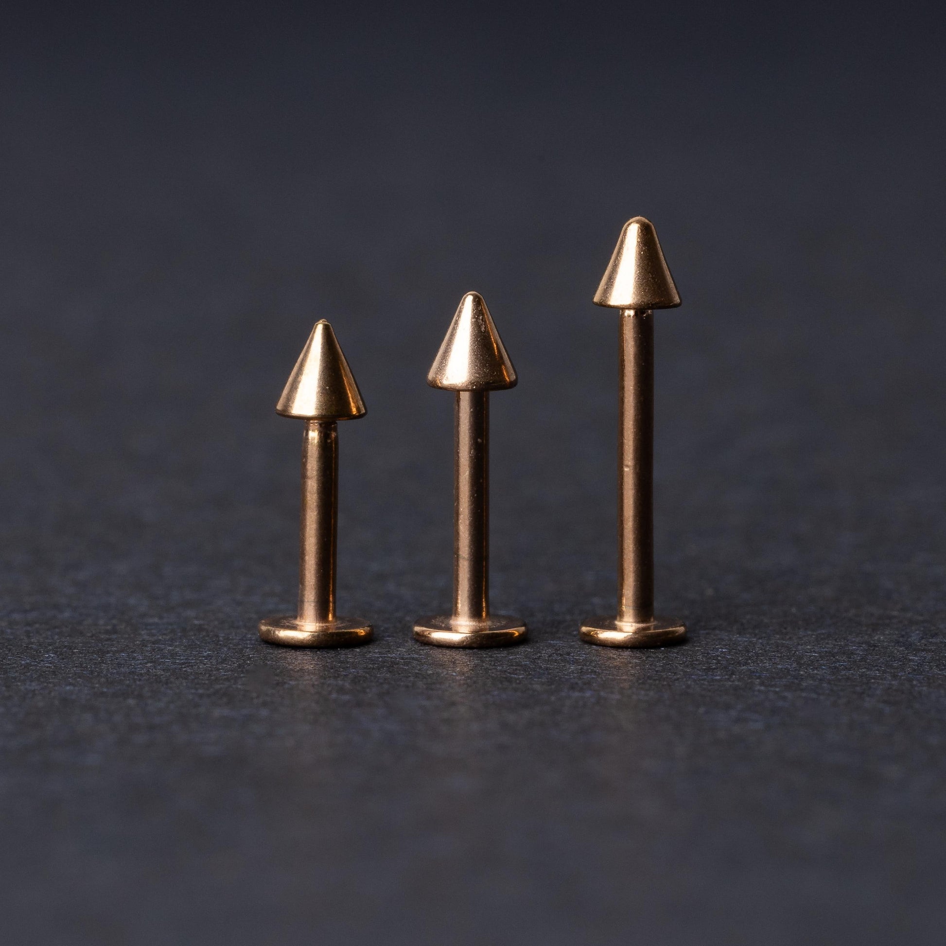 Three rose gold-tone stainless steel labrets with spike ends, standing upright on a dark background.