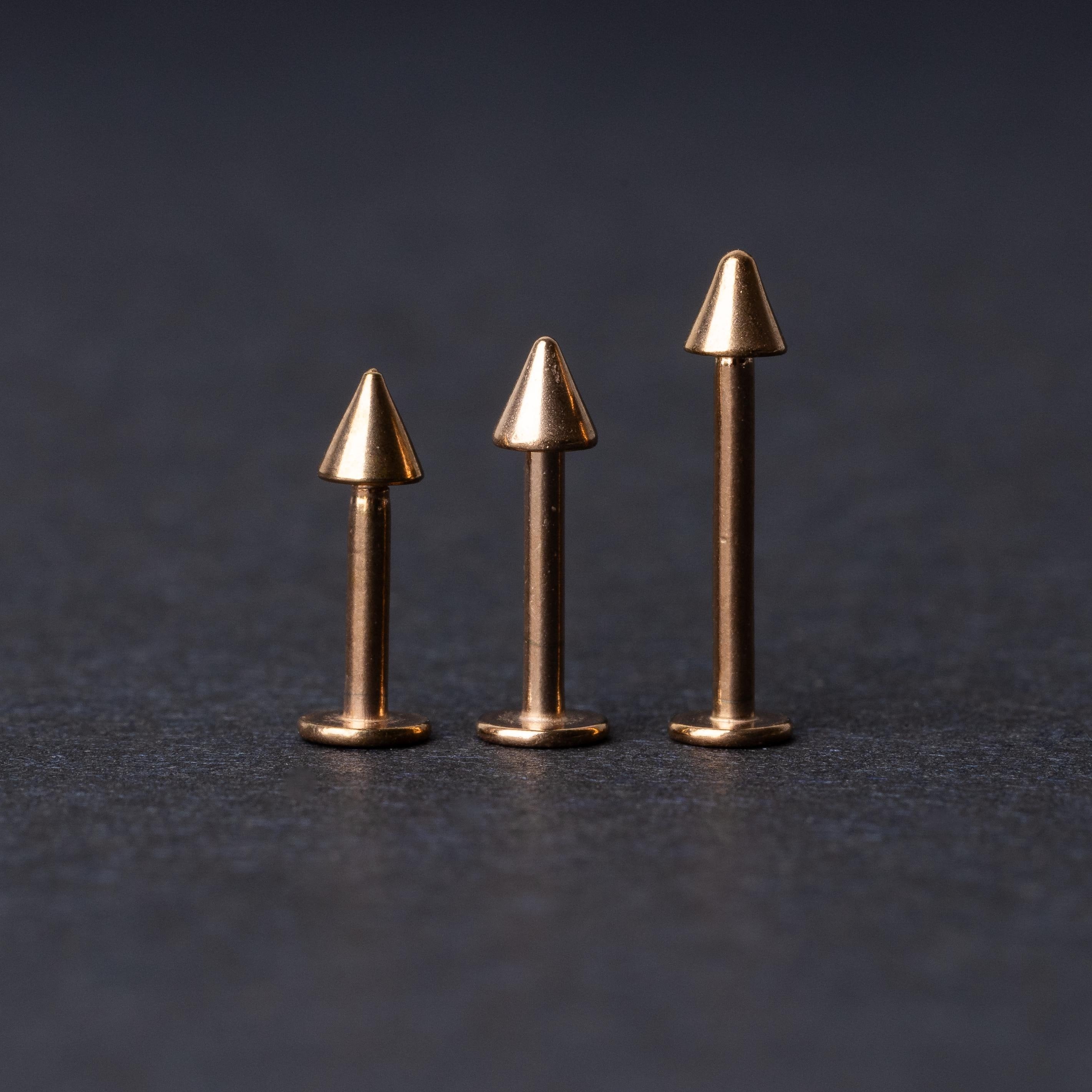 Three rose gold-tone stainless steel labrets with spike ends, standing upright on a dark background.