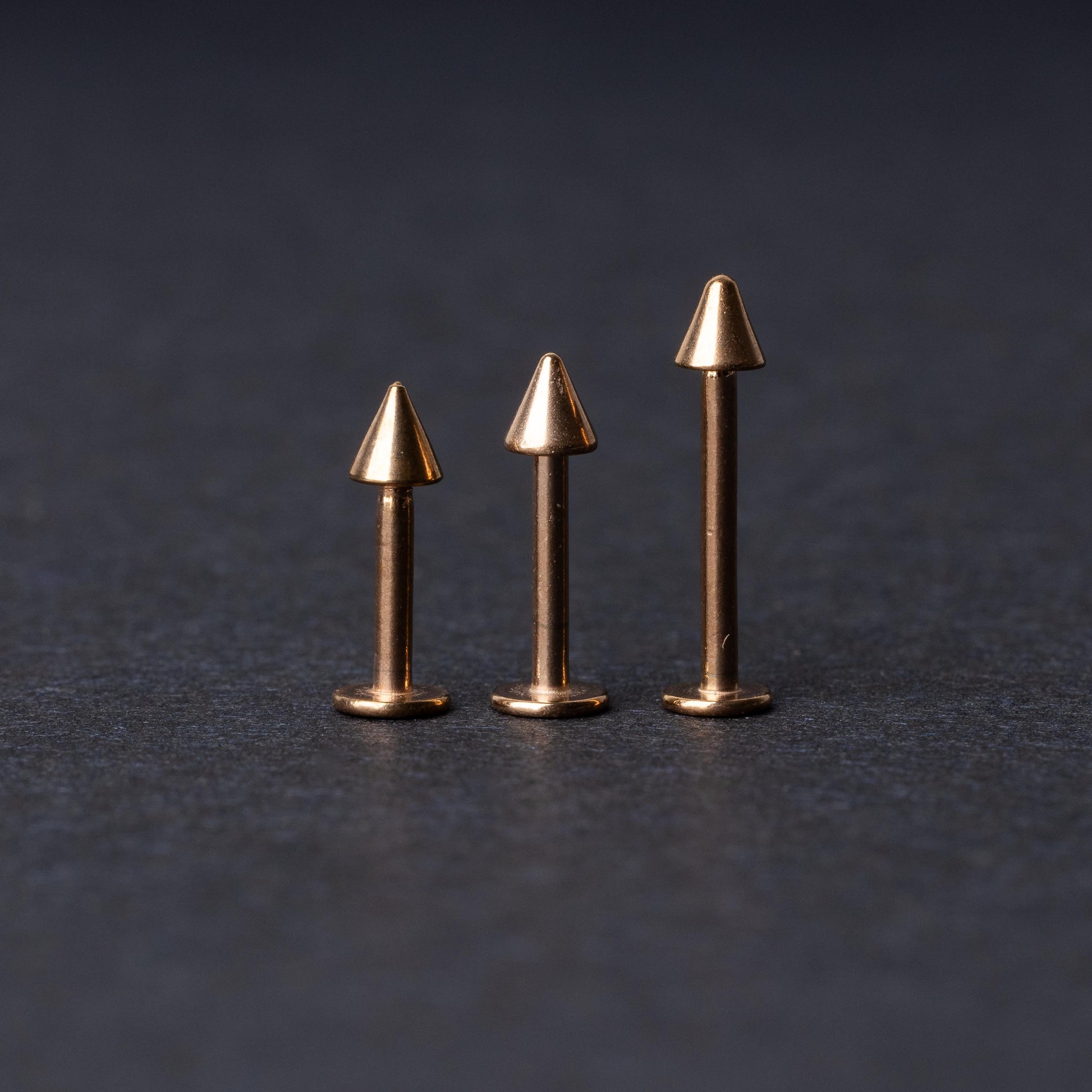 Three rose gold-tone stainless steel labrets with spike ends, standing upright on a dark background.