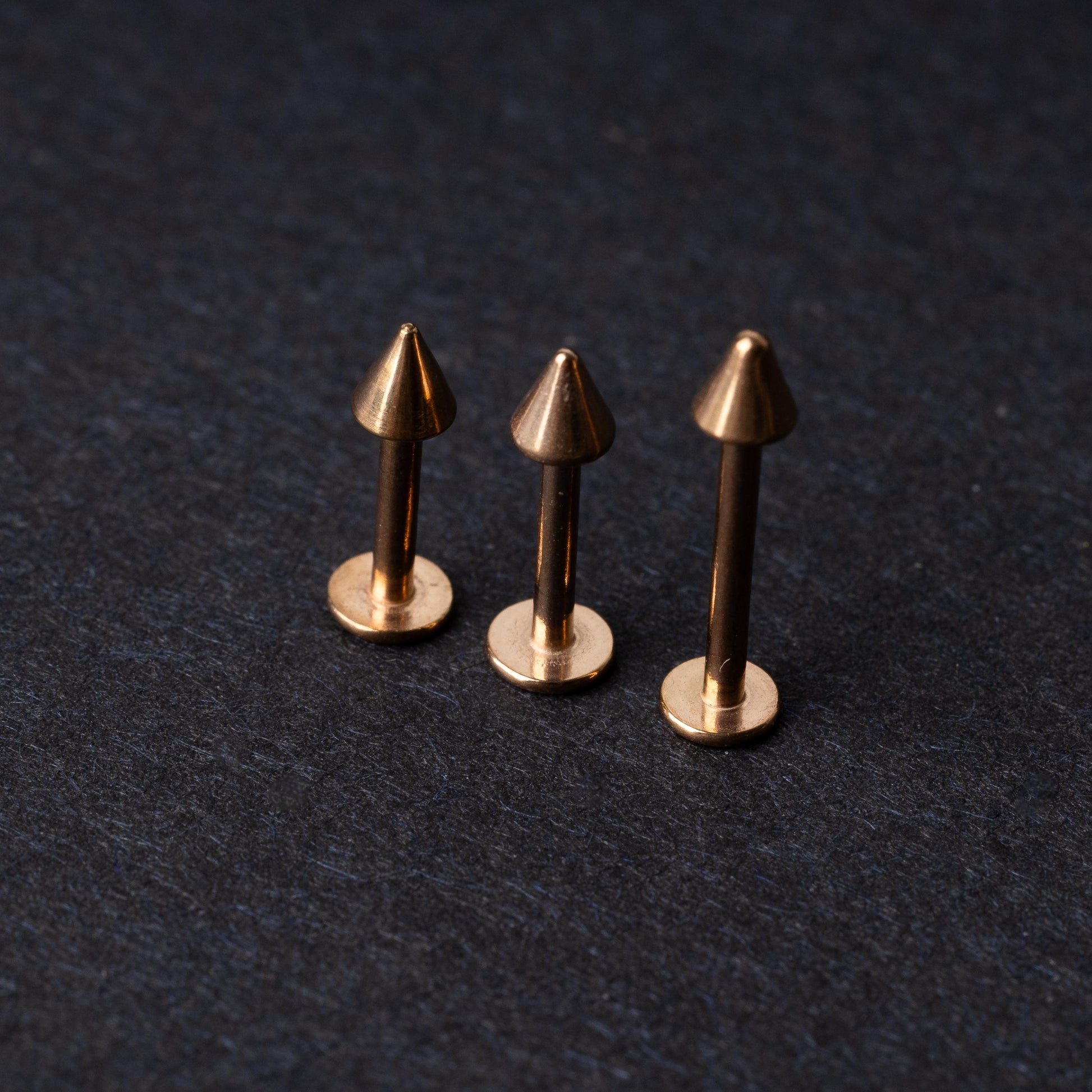 Three rose gold-tone stainless steel labrets with spike ends, standing upright on a dark background.