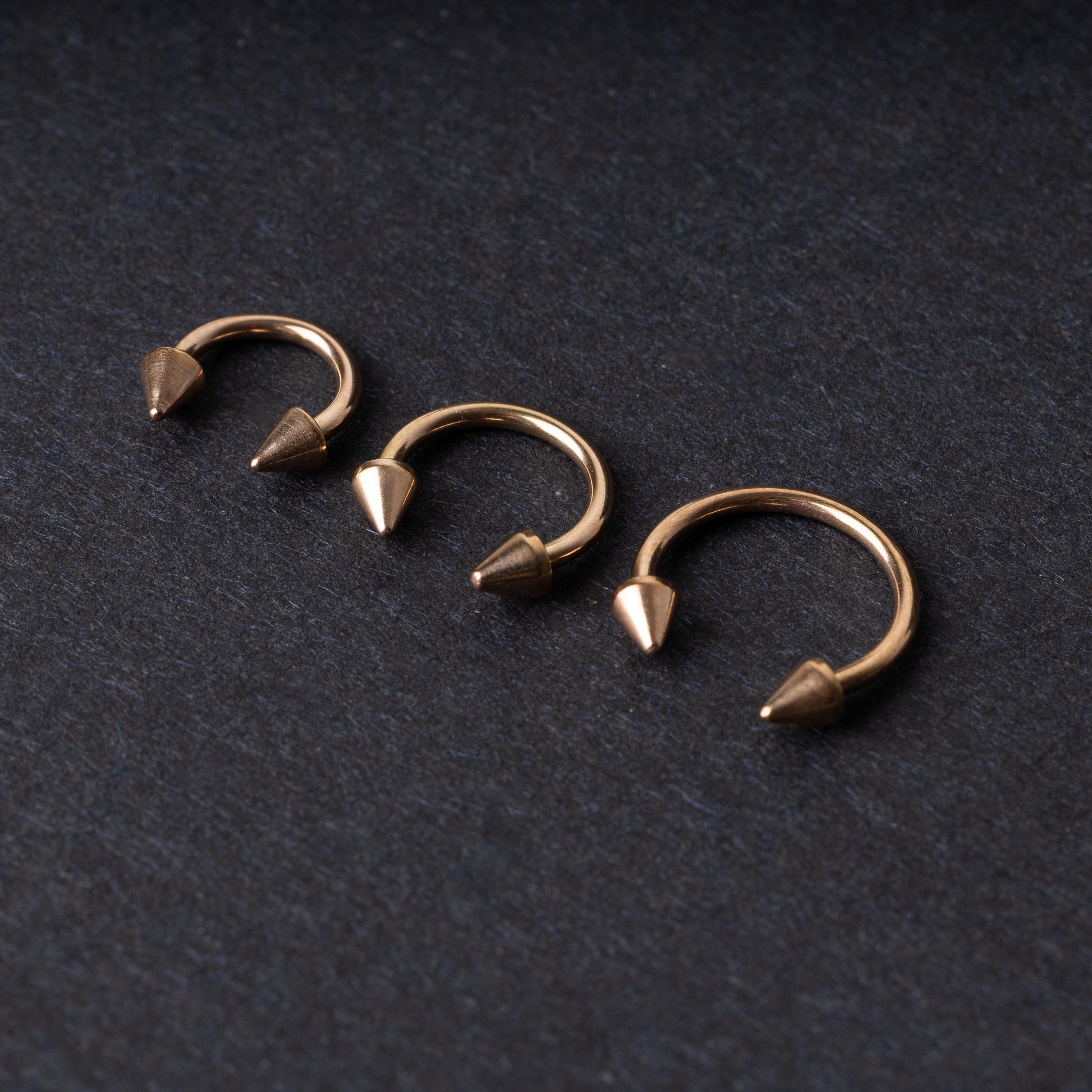 Three rose gold-tone horseshoe hoops with threaded spike ends arranged on a dark textured background.