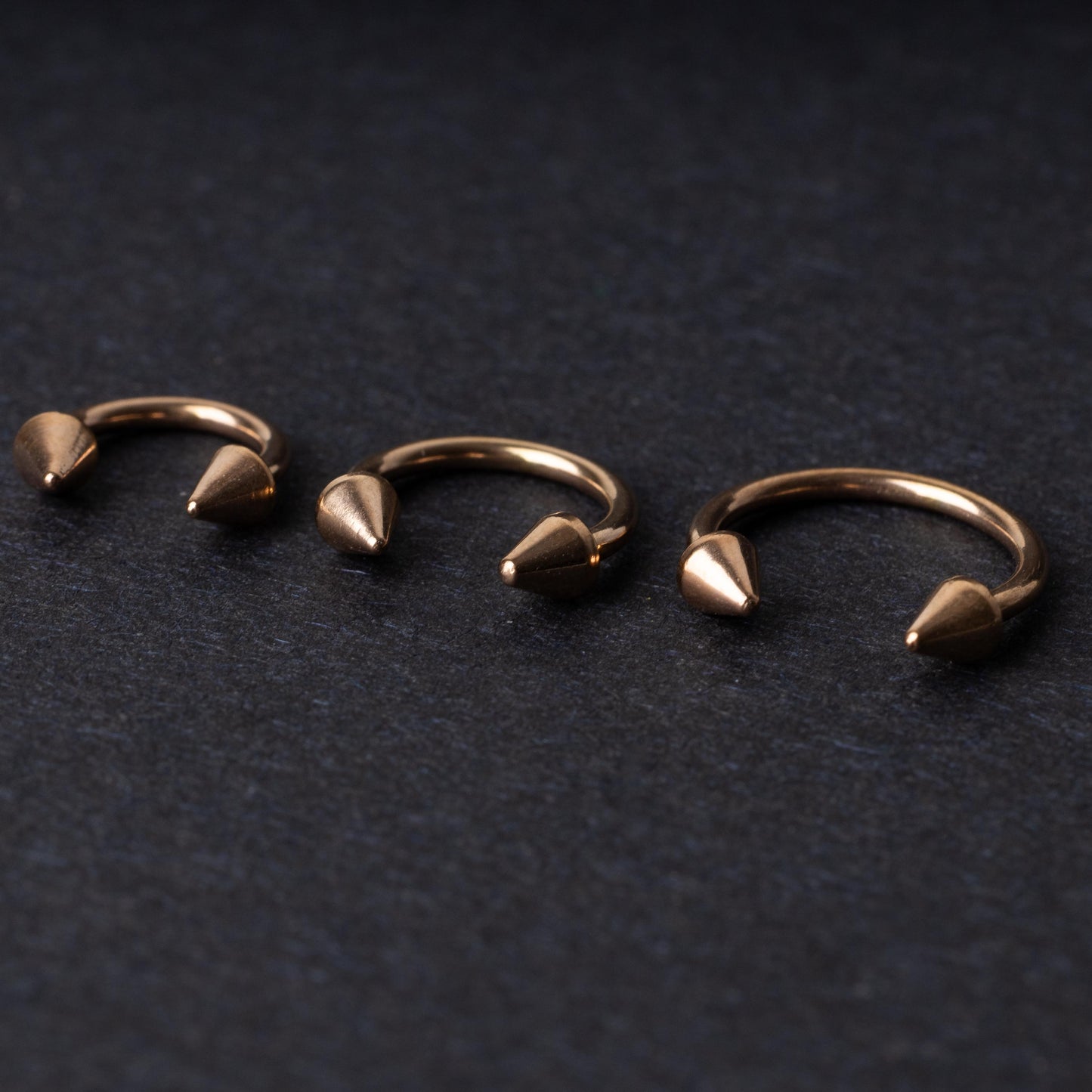 Three rose gold-tone horseshoe hoops with threaded spike ends arranged on a dark textured background.
