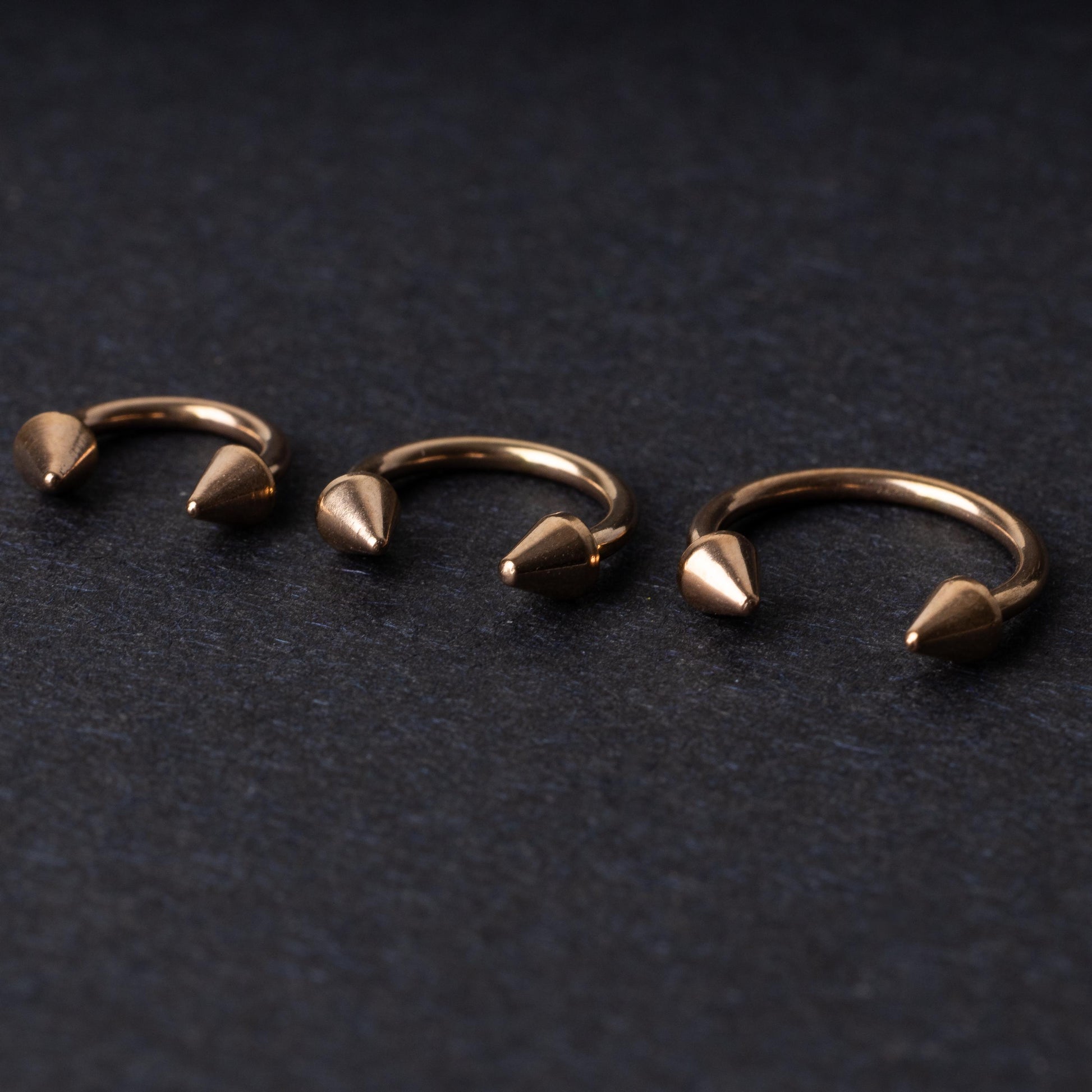 Three rose gold-tone horseshoe hoops with threaded spike ends arranged on a dark textured background.