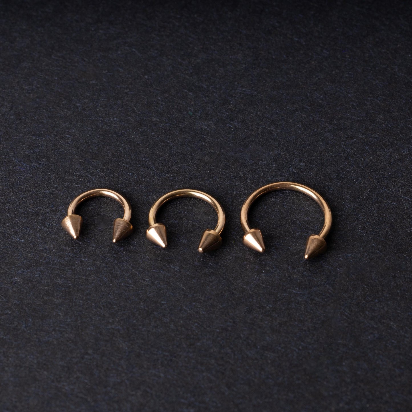 Three rose gold-tone horseshoe hoops with threaded spike ends arranged on a dark textured background.