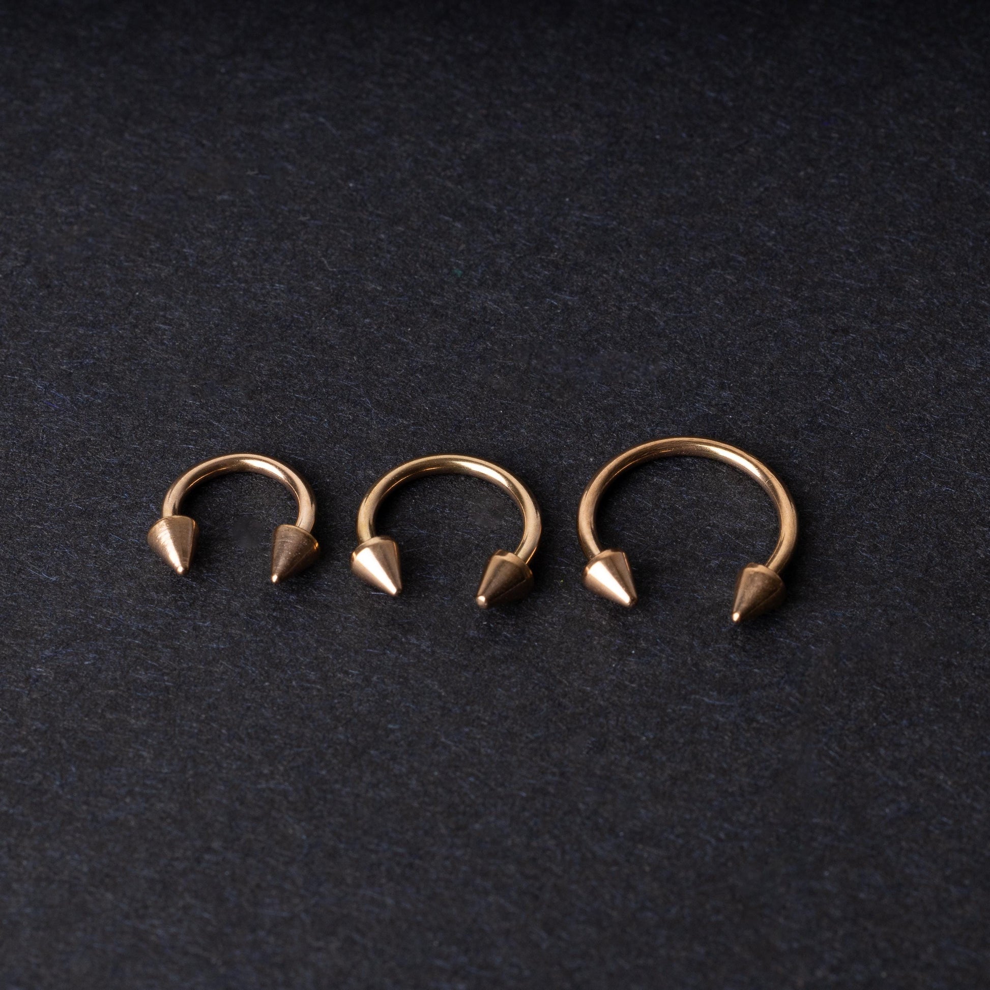 Three rose gold-tone horseshoe hoops with threaded spike ends arranged on a dark textured background.