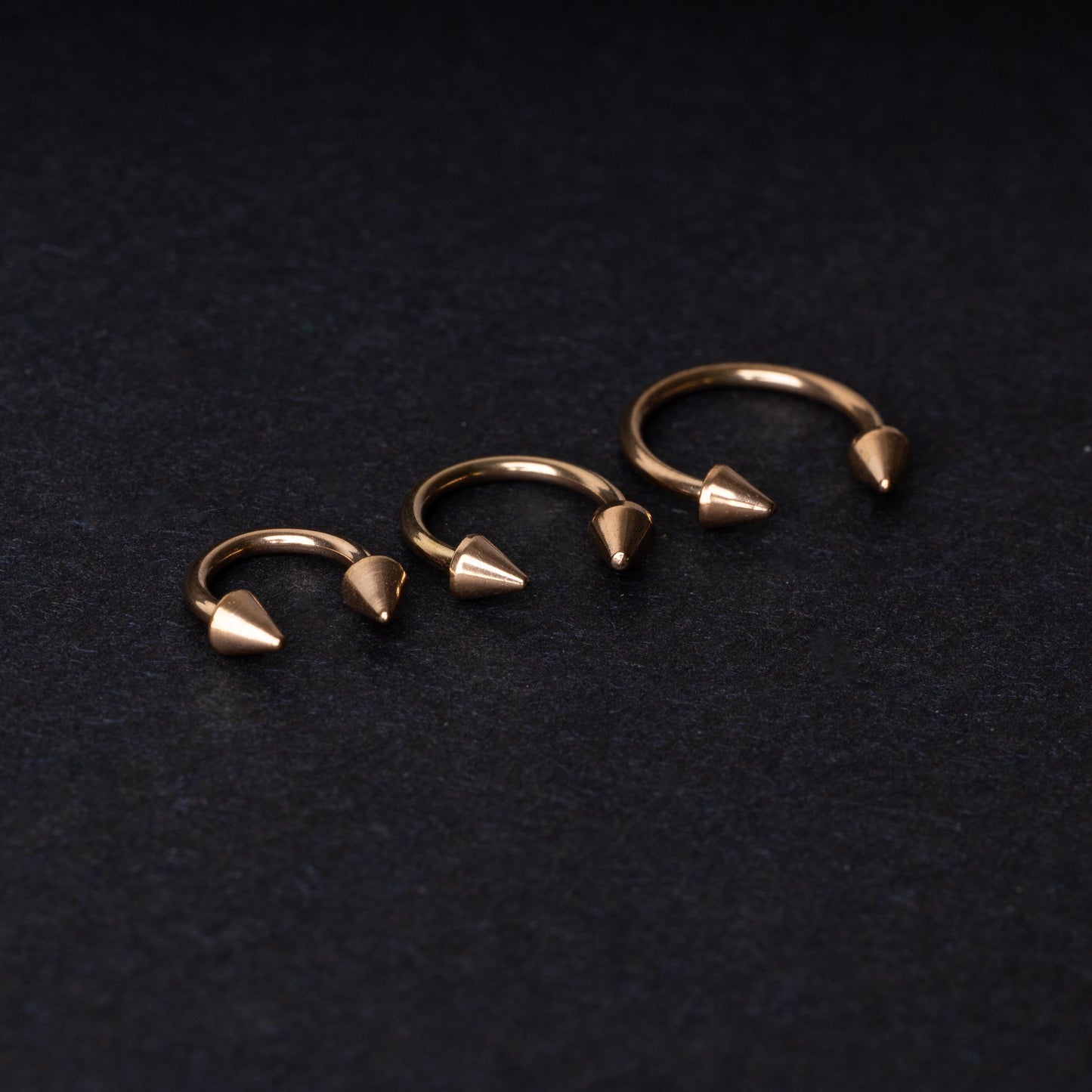 Three rose gold-tone horseshoe hoops with threaded spike ends arranged on a dark textured background.
