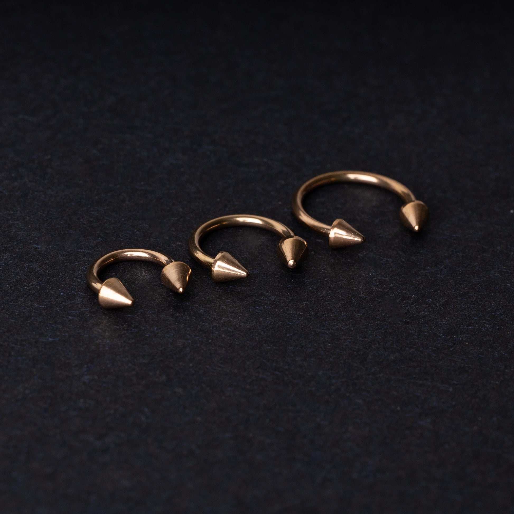 Three rose gold-tone horseshoe hoops with threaded spike ends arranged on a dark textured background.